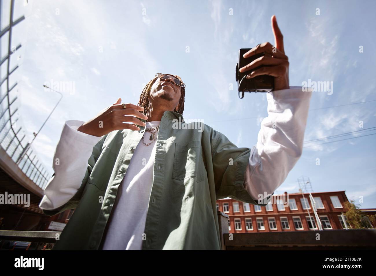 Ammira dal basso il giovane afroamericano con walkman ® in mano che esegue hip hop contro il cielo blu in un ambiente urbano Foto Stock