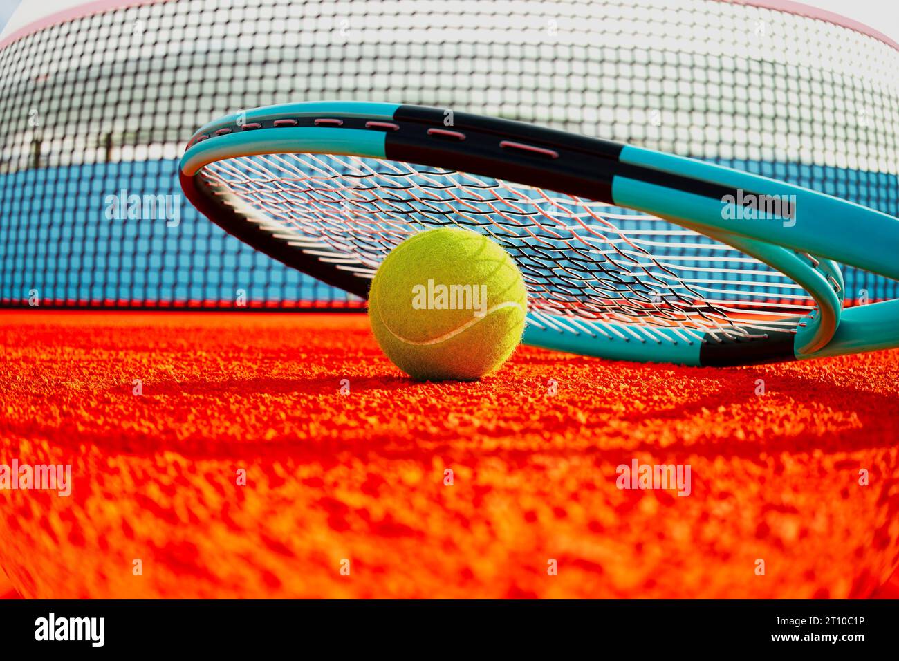 Una prospettiva ravvicinata di una vibrante palla da tennis gialla che poggia su un campo arancione, protetto da una racchetta da tennis, con uno sfondo di una rete. Dinamica e. Foto Stock