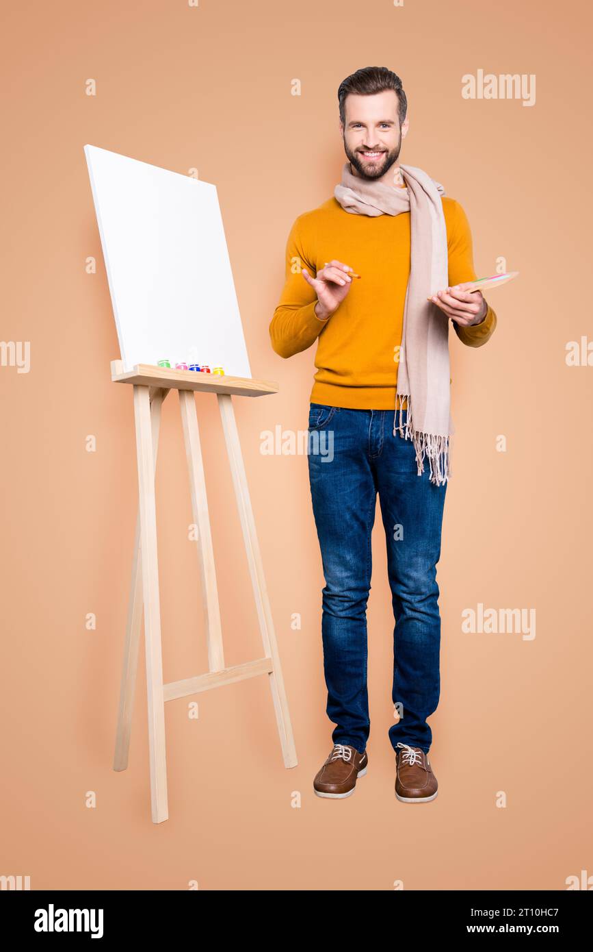 Ritratto integrale di gioioso artista trendy con sciarpa intorno al collo, acconciatura, in jeans, maglione, con palette colorate e pennelli in mano Foto Stock