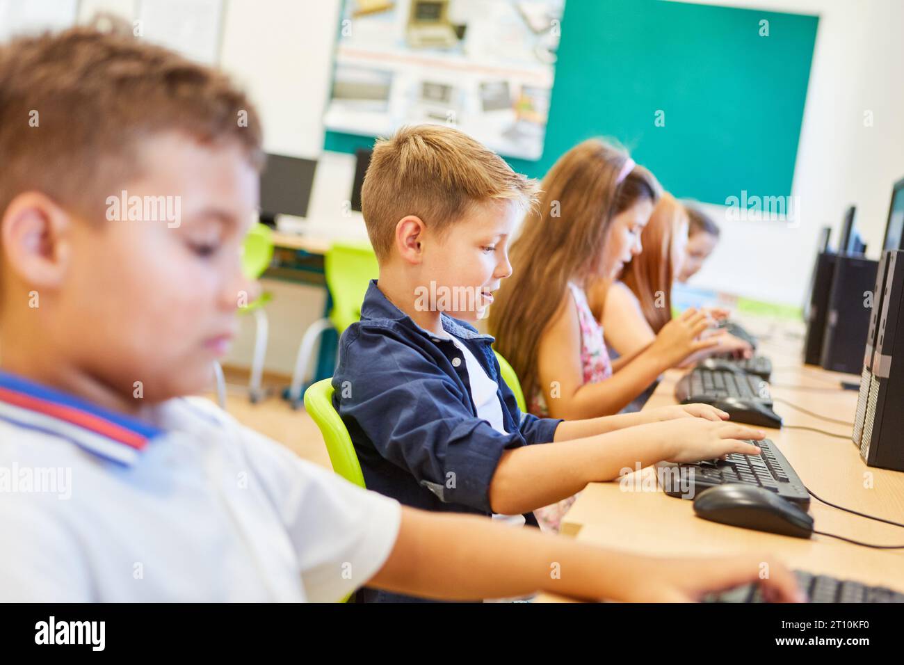 Studente elementare che digita sulla tastiera mentre si siede con gli amici in una lezione di computer Foto Stock
