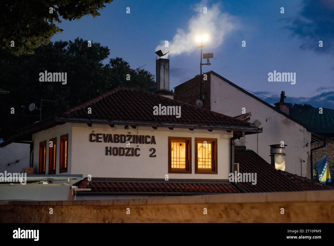 Esplora l'atmosfera accogliente di un ristorante Cevape a Sarajevo, in Bosnia, mentre cala la notte. Le luci calde si illuminano e il camino emette vapore. Foto Stock