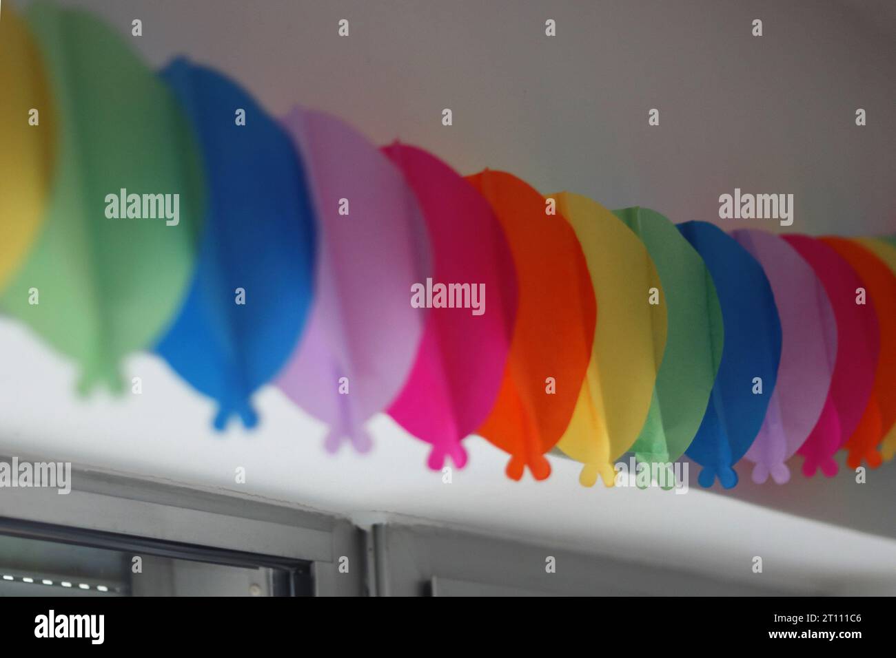 Colorata ghirlanda di compleanno sulla parete bianca sopra la finestra. Facciamo festa. Decorazione murale di compleanno. Catena di carta decorata per le festività sulla parete bianca abov Foto Stock