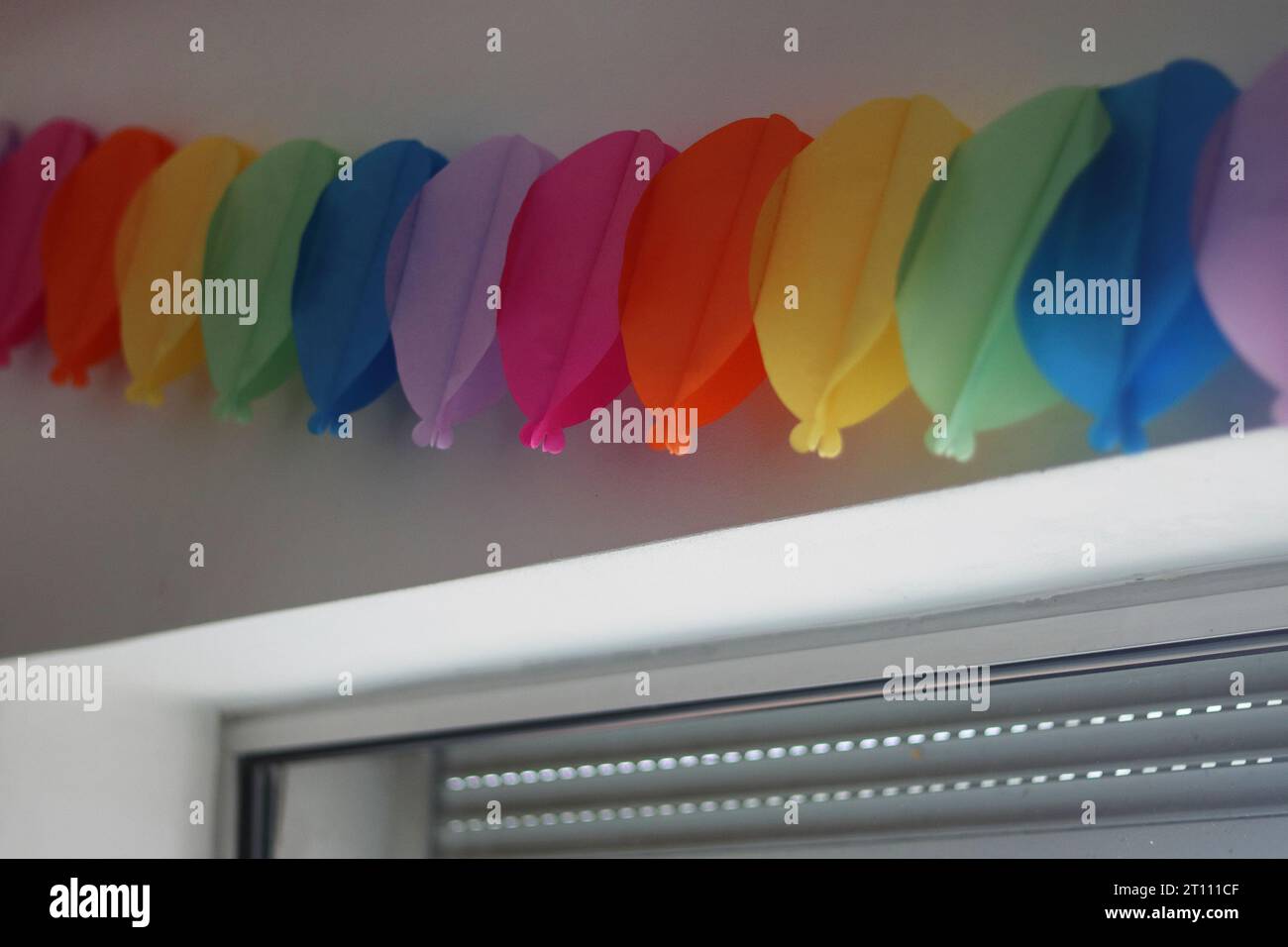 Colorata ghirlanda di compleanno sulla parete bianca sopra la finestra. Facciamo festa. Decorazione murale di compleanno. Catena di carta decorata per le festività sulla parete bianca abov Foto Stock