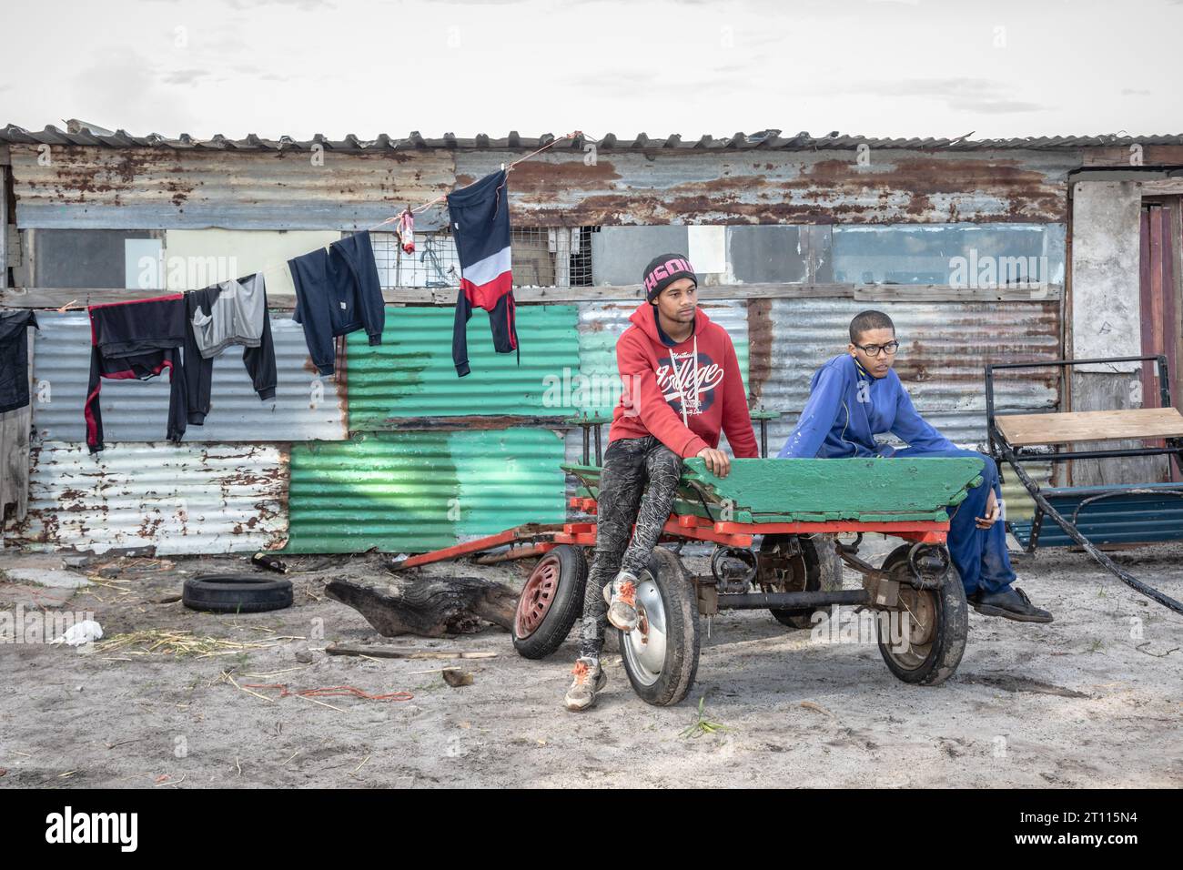 Due giovani siedono su un carro davanti a una baracca di ferro ondulato in una povera comunità in Sudafrica Foto Stock