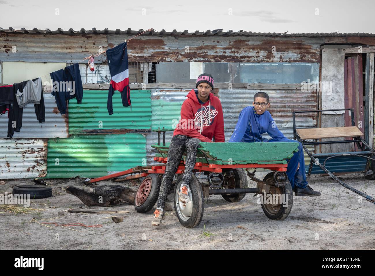 Due giovani siedono su un carro davanti a una baracca di ferro ondulato in una povera comunità in Sudafrica Foto Stock