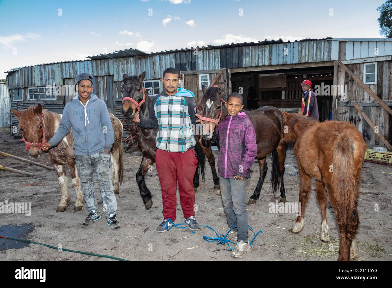 Due giovani uomini e bambini di razza mista in piedi accanto ai loro cavalli da carro, usati per trainare carri per trasportare merci nella comunità povera del Sudafrica Foto Stock