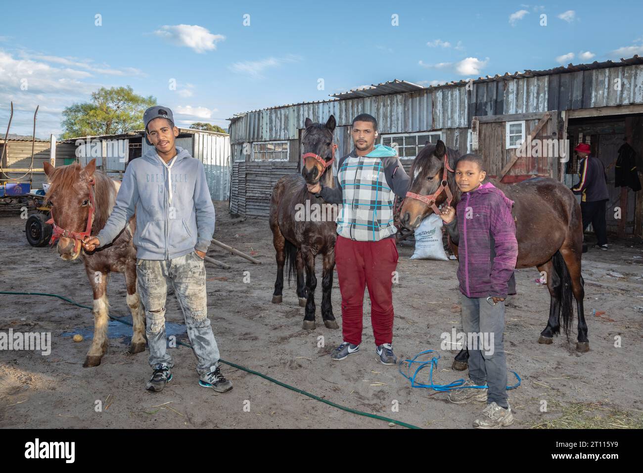 Due giovani uomini e bambini di razza mista in piedi accanto ai loro cavalli da carro, usati per trainare carri per trasportare merci nella comunità povera del Sudafrica Foto Stock