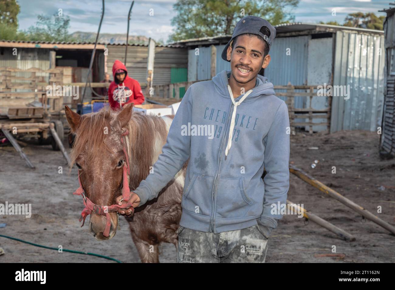 Un giovane uomo di razza mista che si trova accanto al suo cavallo da carro, usato per trainare carrelli per trasportare merci nella comunità povera del Sudafrica Foto Stock