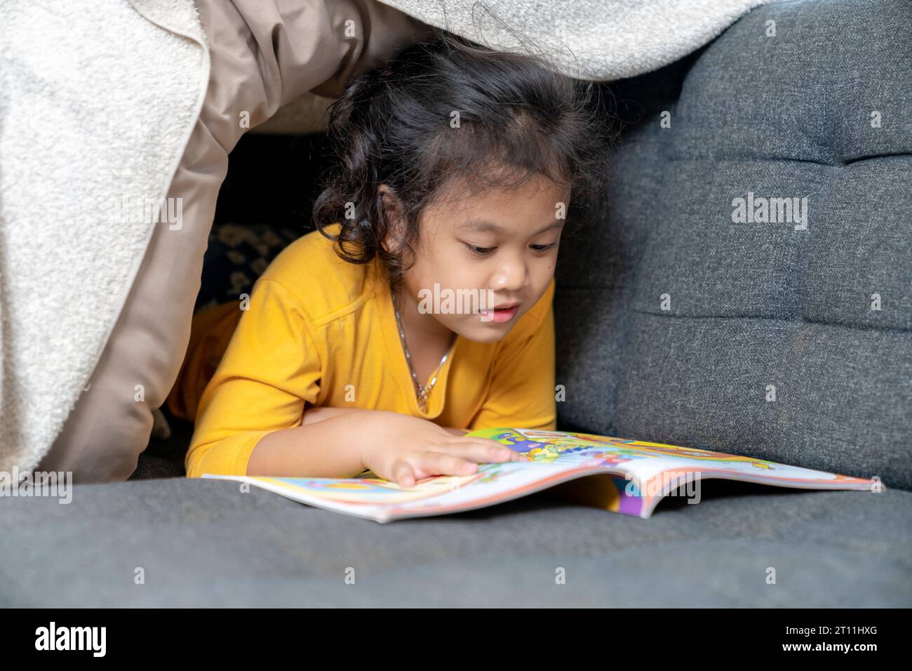 Piccola e carina ragazza asiatica riccia che legge un libro in tenda da gioco a casa, bambino felice che gioca. Foto Stock