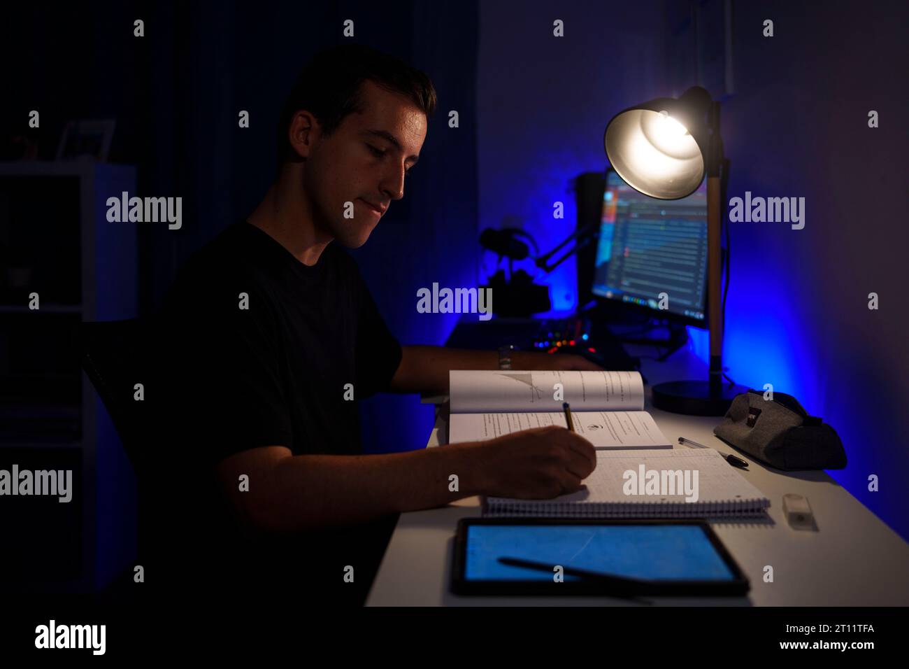 Vista laterale di un giovane che prende appunti mentre studia nella sua camera da letto, illuminato da una lampada da scrivania Foto Stock