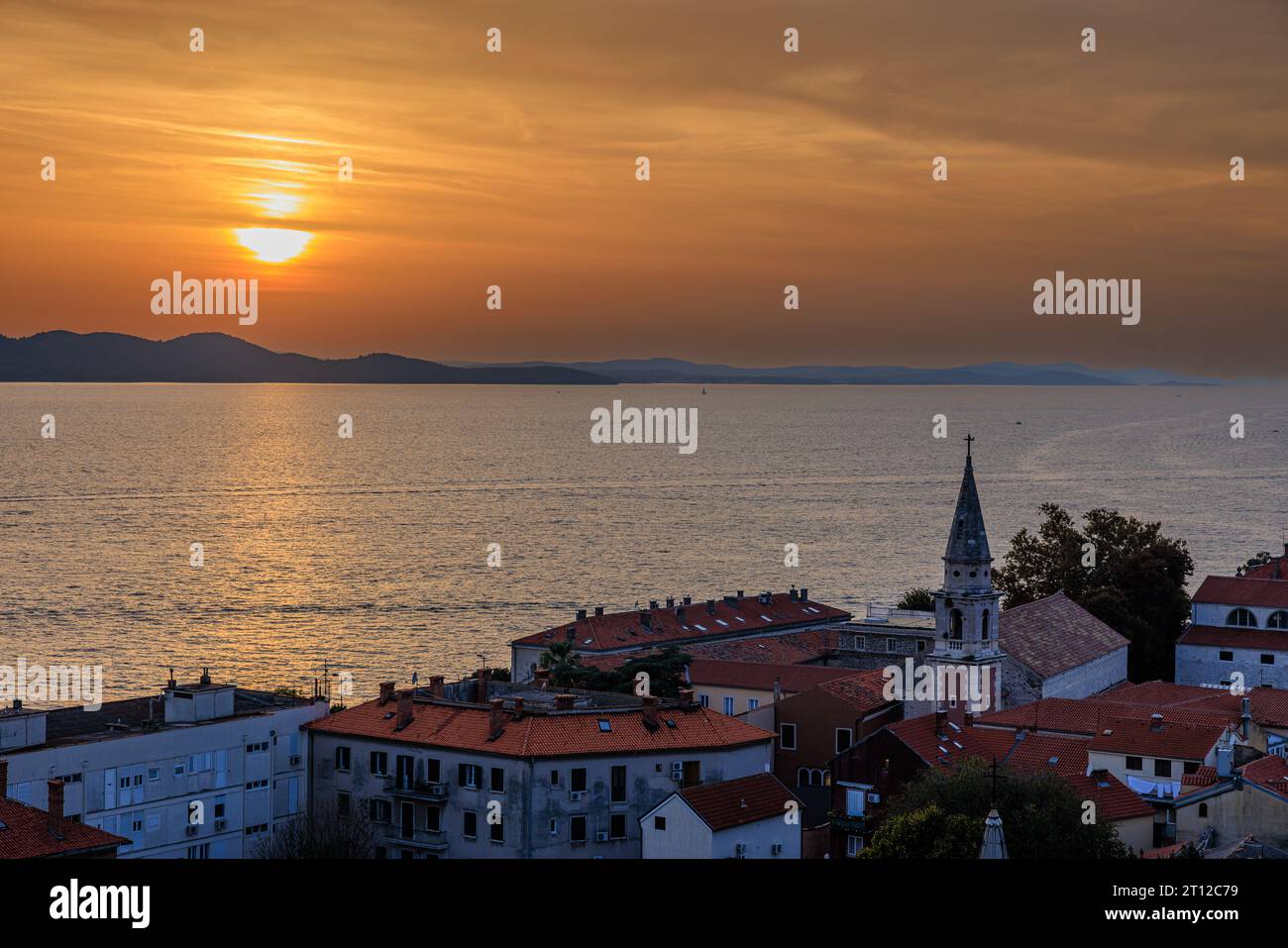 vista aerea del sole che tramonta dietro montagne lontane in un cielo arancione riflesso in un mare calmo dal campanile di sant'anastasia a zara in croazia Foto Stock