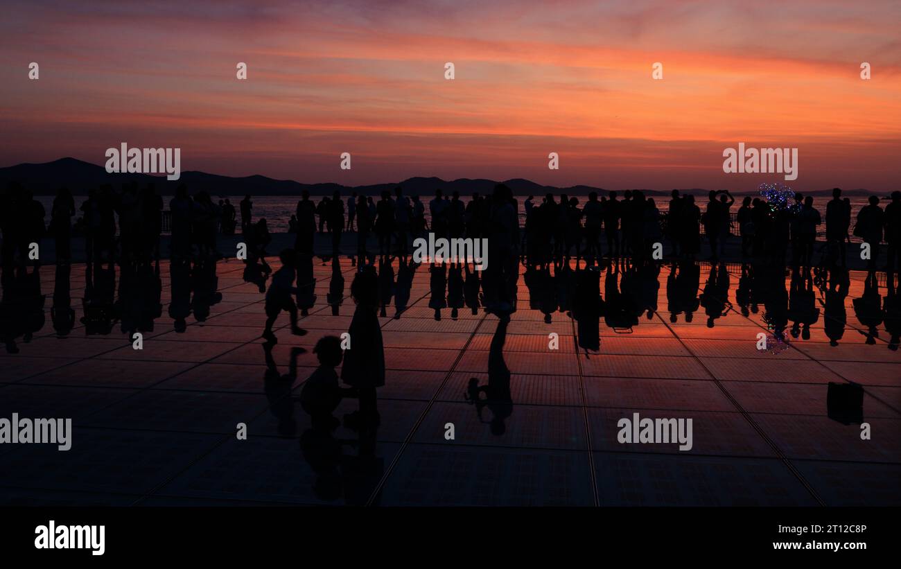 la folla si riunisce intorno a un sassofonista solista al tramonto riflesso nei pannelli solari del saluto all'installazione del sole al tramonto a zara in croazia Foto Stock