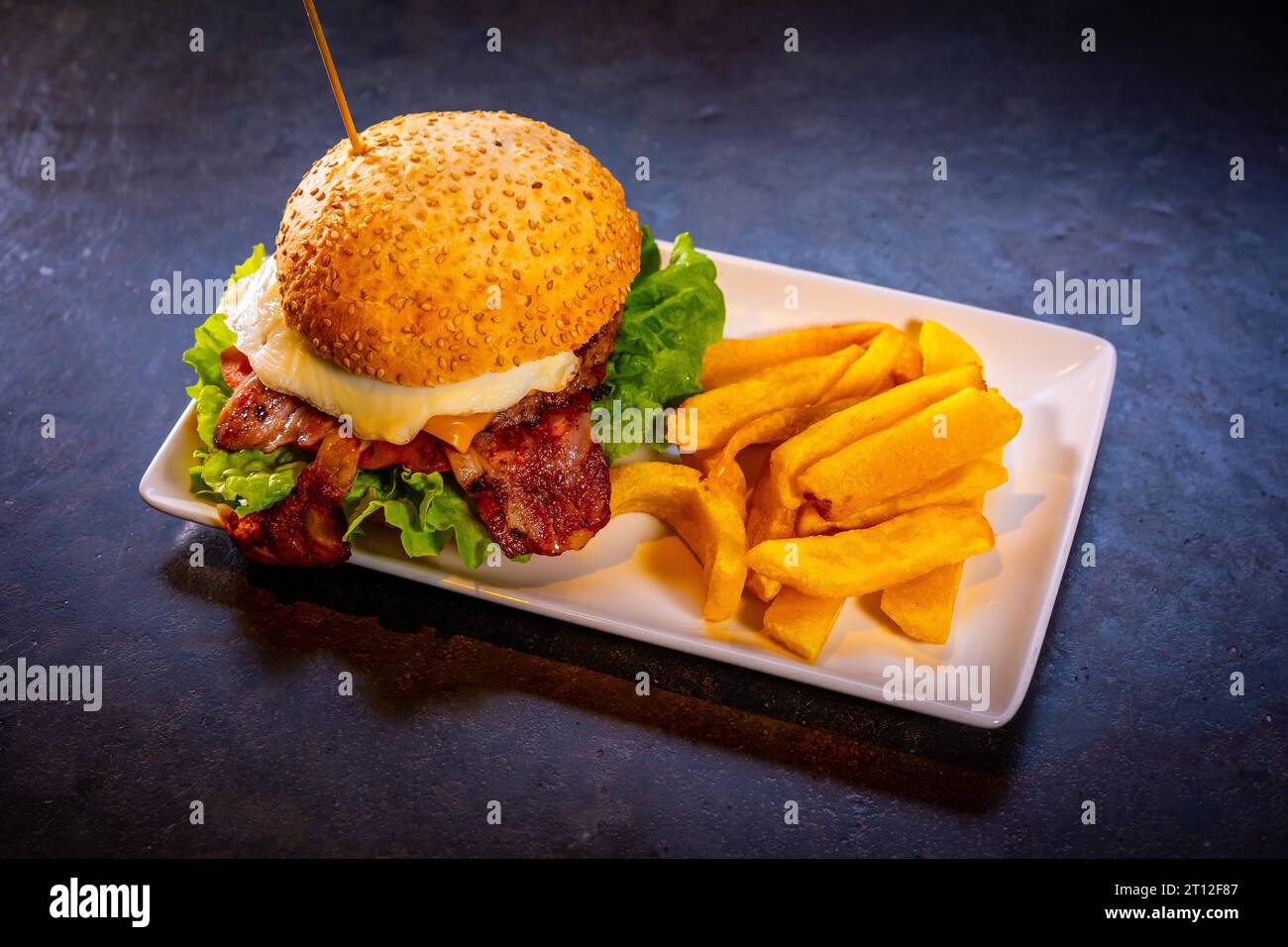 Hamburger con pancetta e patatine fritte su fondo nero, su un piatto bianco Foto Stock