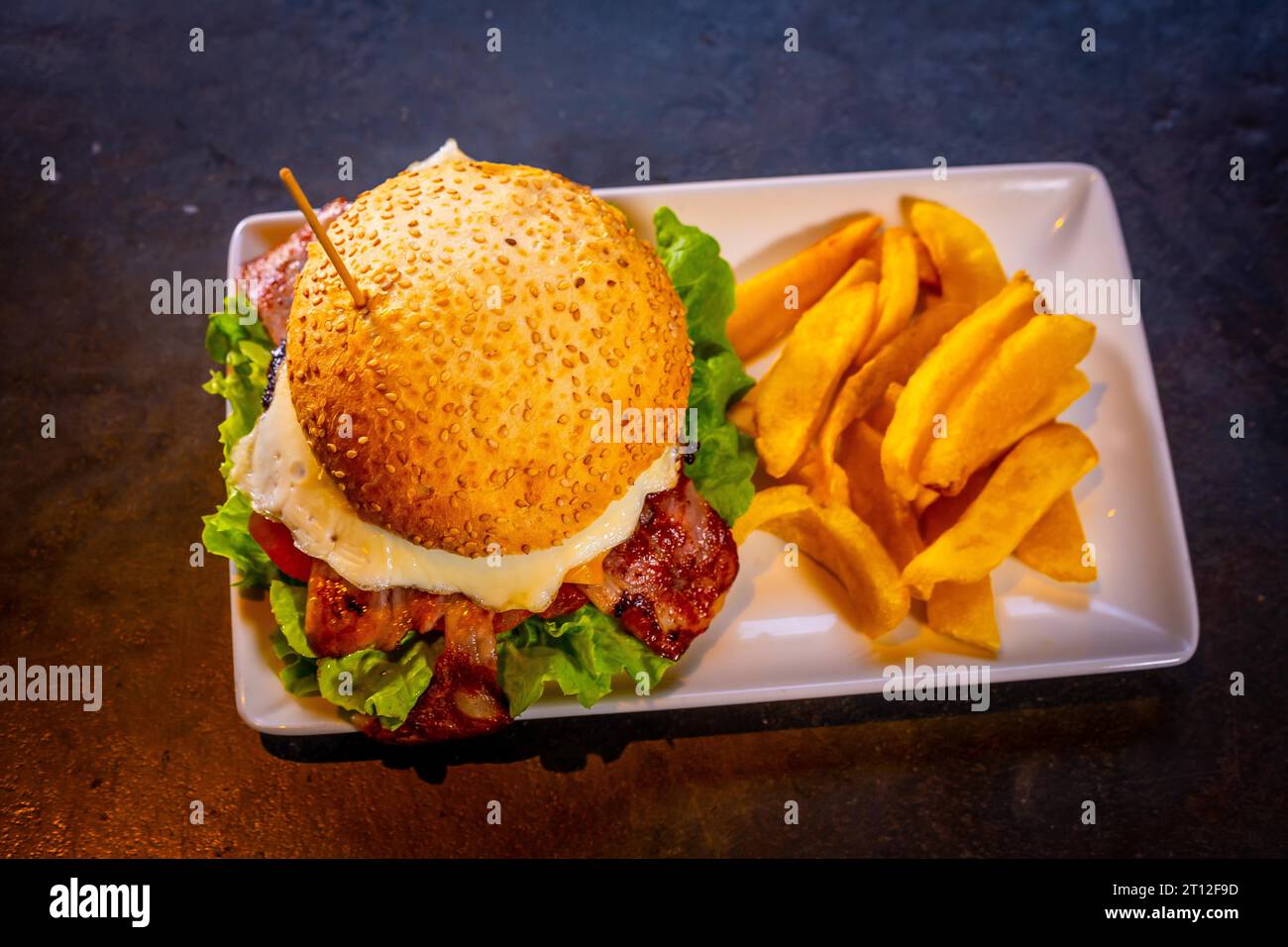 Hamburger con pancetta e patatine fritte su fondo nero, su un piatto bianco Foto Stock