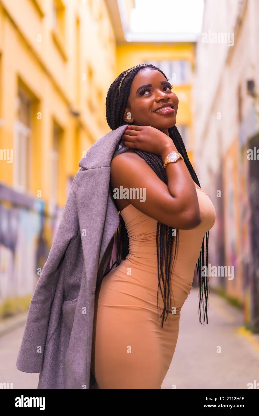 Stile urbano alla moda con una ragazza africana nera in un abito color crema su una strada cittadina. In posa con una giacca sulla spalla Foto Stock