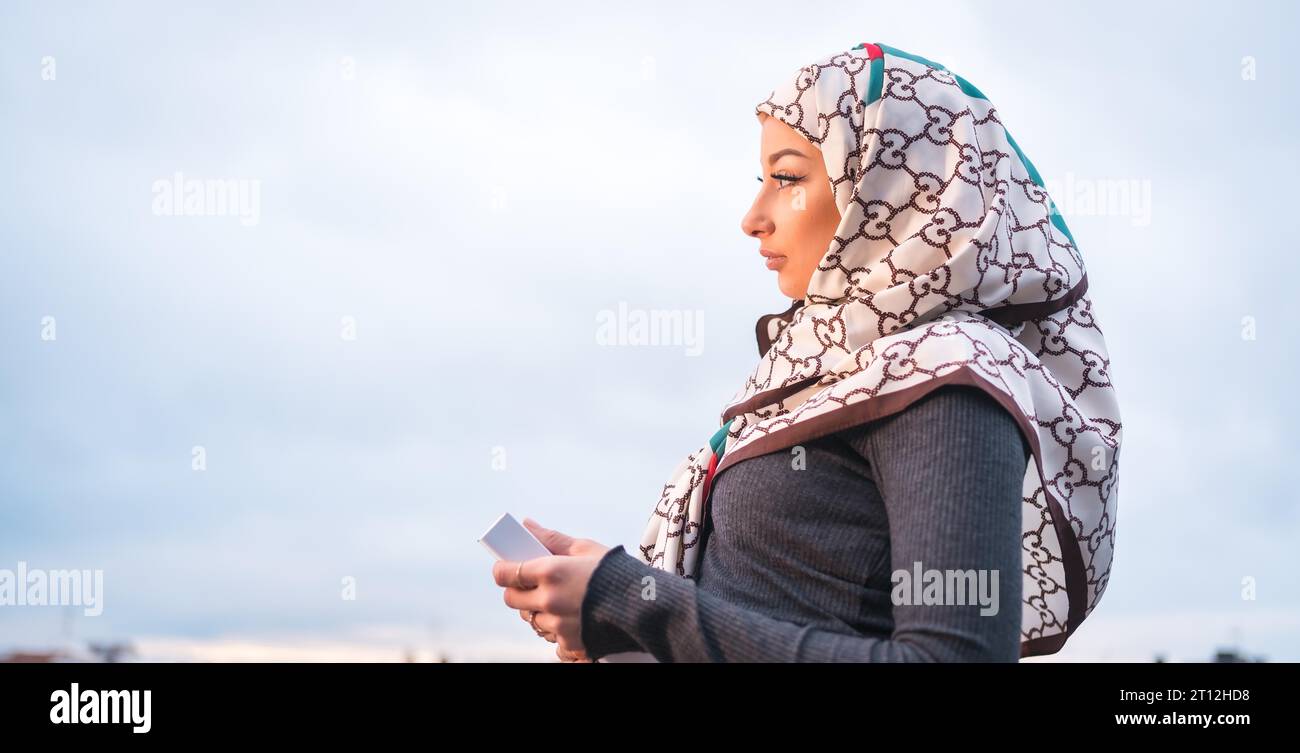 Ritratto di una ragazza araba con un velo bianco con un libro sulla terrazza di una caffetteria, che guarda la città da un balcone Foto Stock