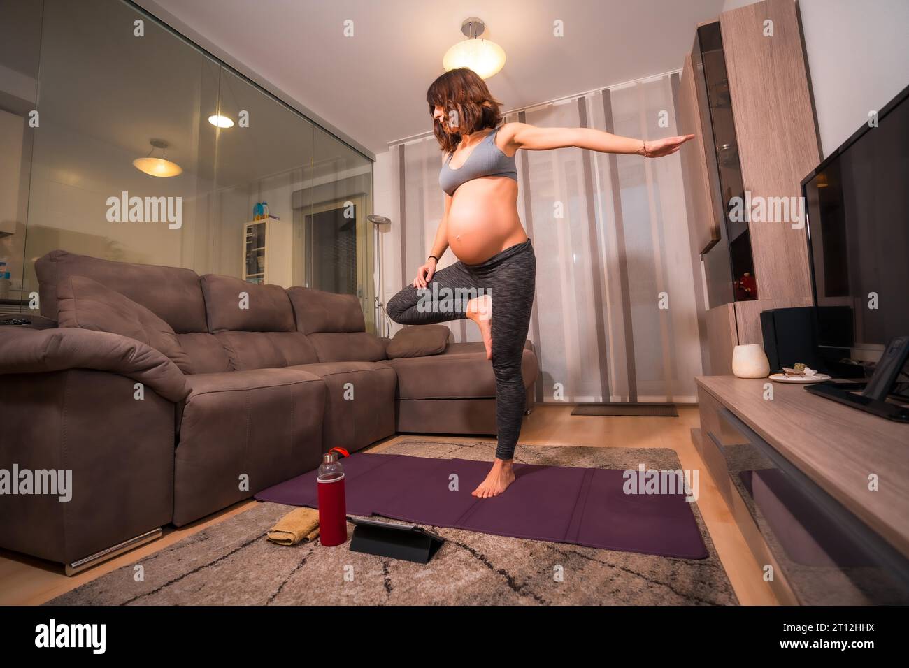 Una giovane donna caucasica incinta con una maglietta grigia che fa esercizio di pilates a casa. Esercizi online in classi preparatorie Foto Stock