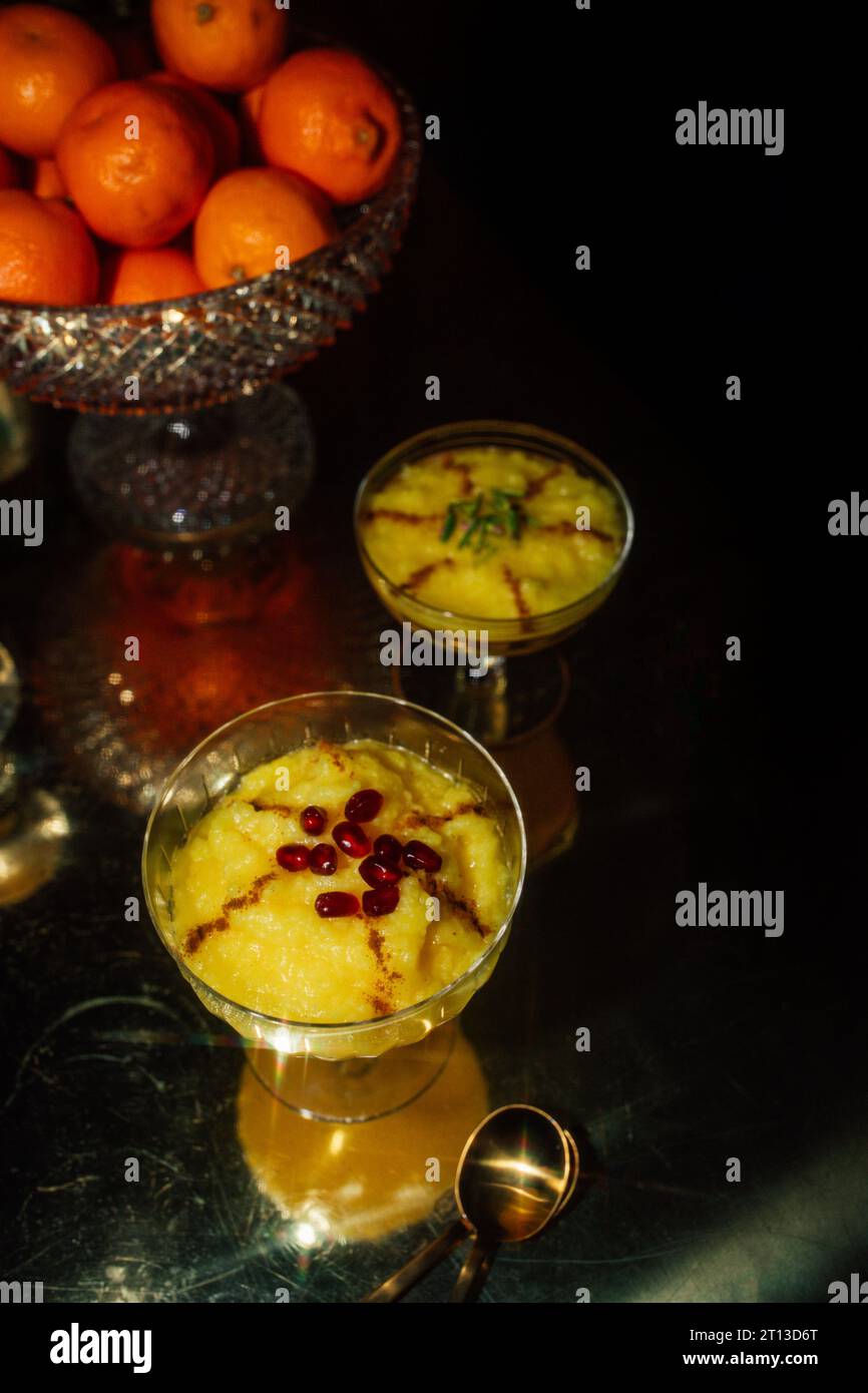 Sholeh Zard, un dessert persiano di riso allo zafferano condito con cannella e pistacchi sul tavolo con una ciotola di frutta di arance e sfondo scuro Foto Stock