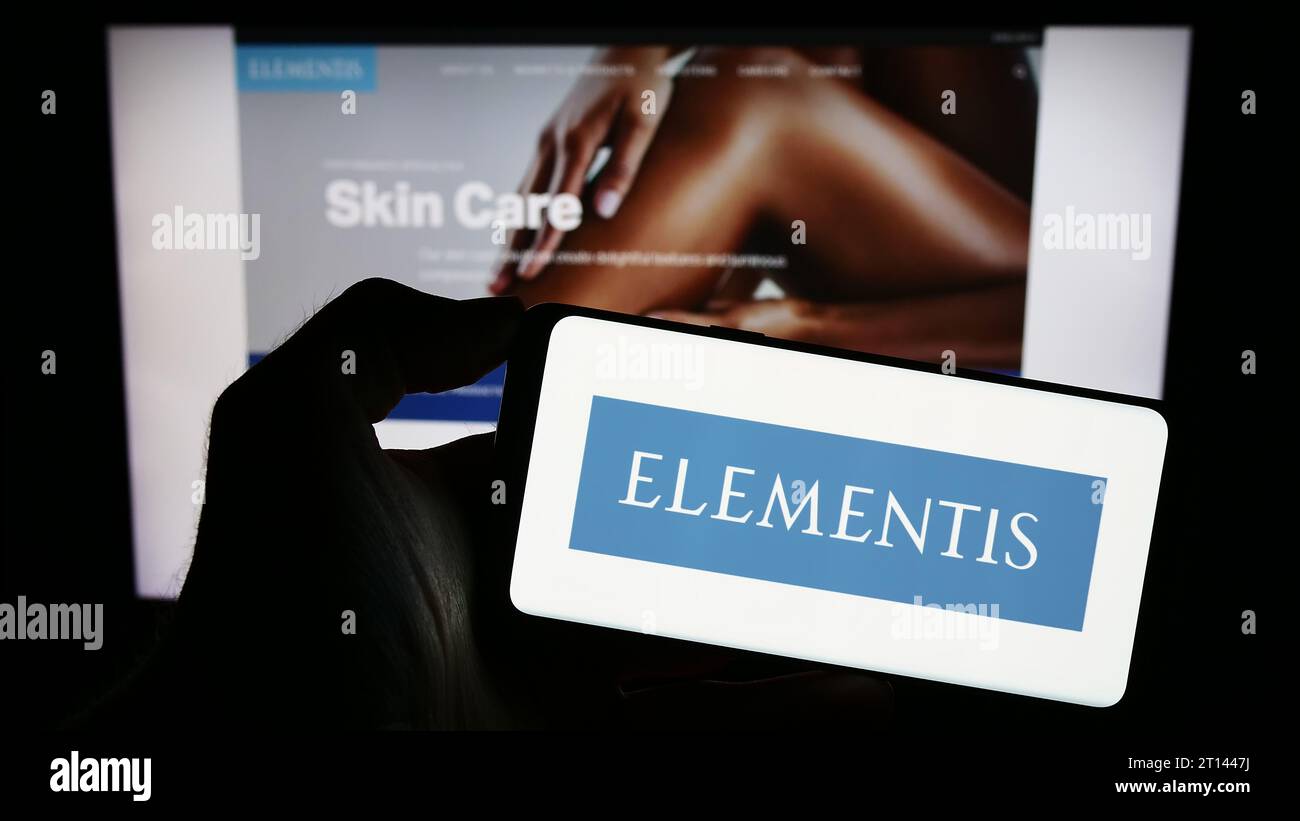 Persona che possiede uno smartphone con il logo dell'azienda britannica Elementis plc di fronte al sito Web. Concentrarsi sul display del telefono. Foto Stock