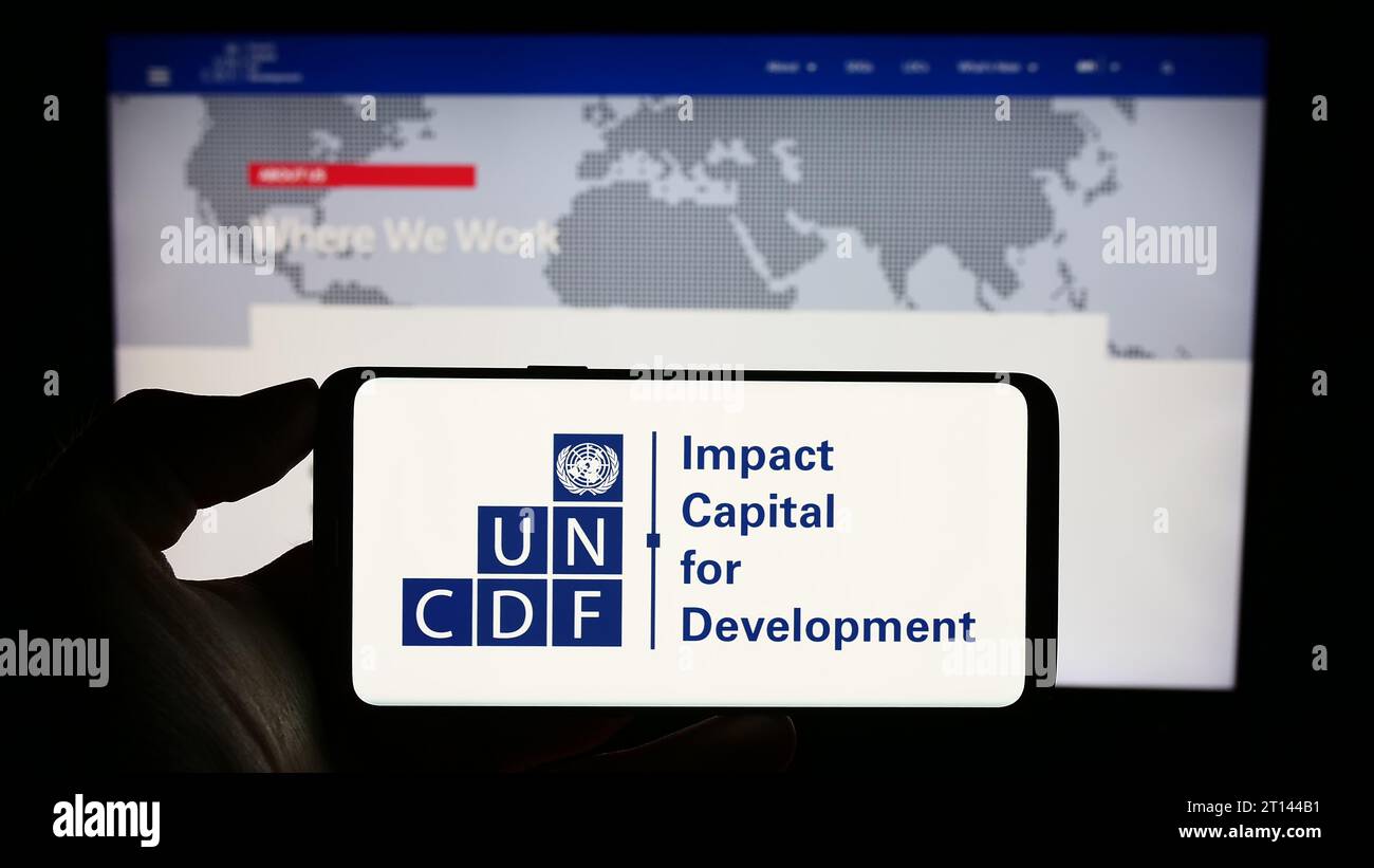 Persona in possesso di un cellulare con il logo del Fondo delle Nazioni Unite per lo sviluppo del capitale (UNCDF) davanti alla pagina web. Concentrarsi sul display del telefono. Foto Stock