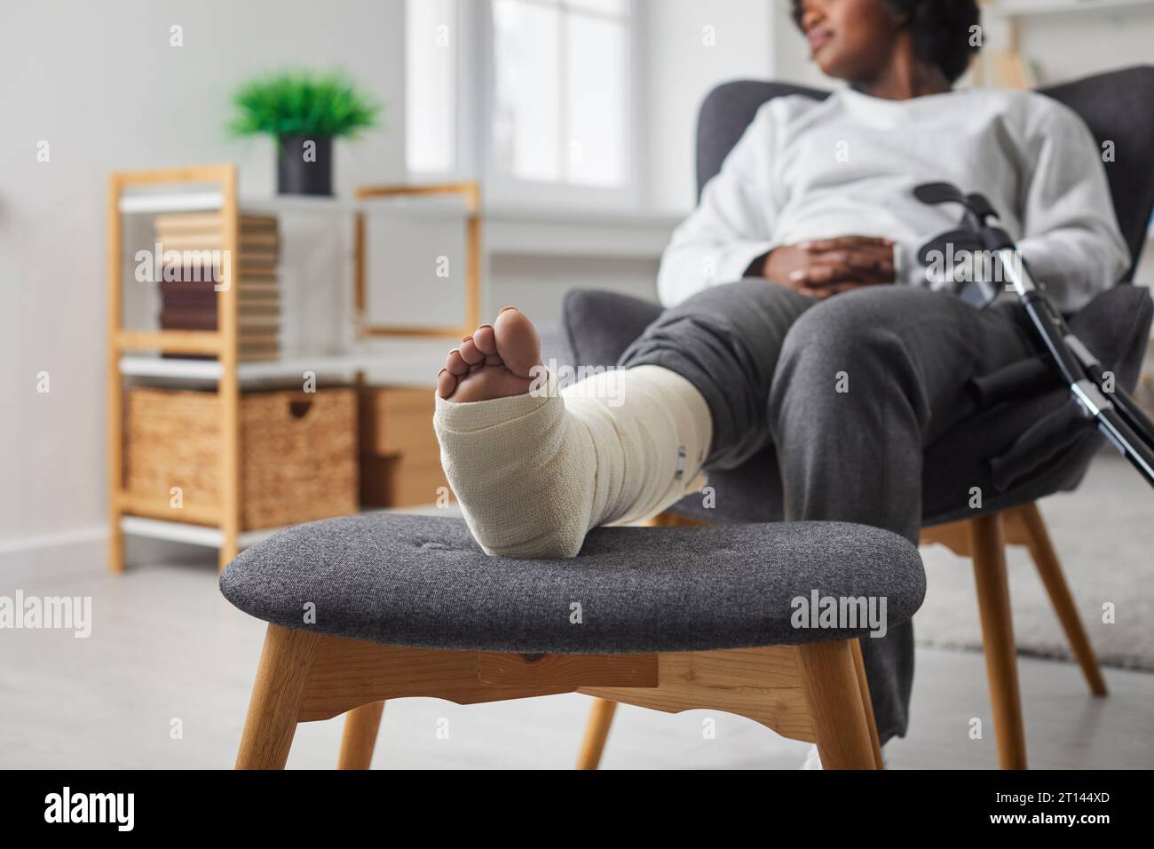 Vista ritagliata di una donna con gamba in gesso fasciatura e poltrona in crutch sittingin a casa Foto Stock