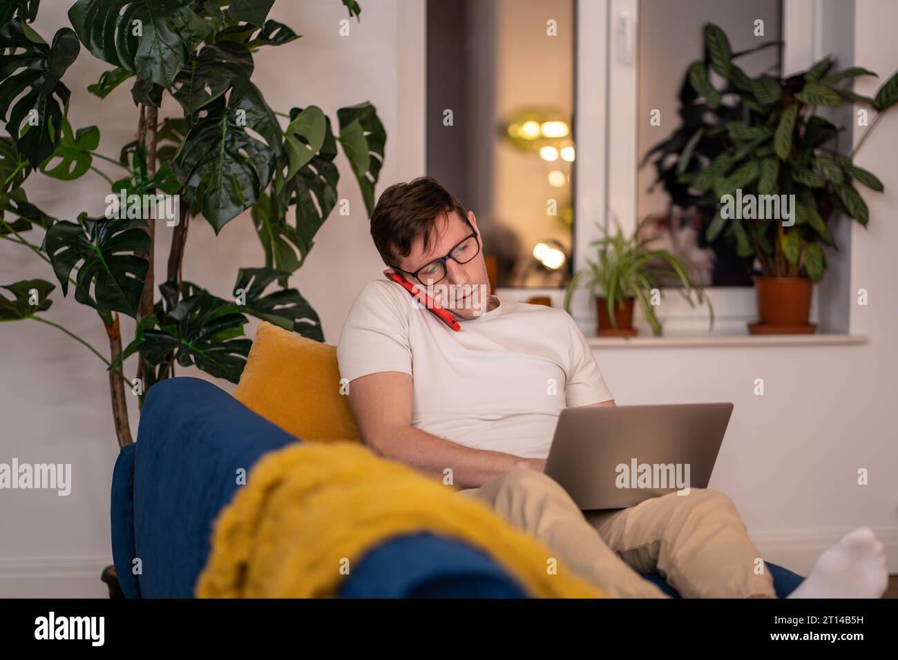 Uomo impegnato seduto sul divano con un computer portatile. Ragazzo serio con occhiali che parla al telefono, concentrato sul lavoro Foto Stock