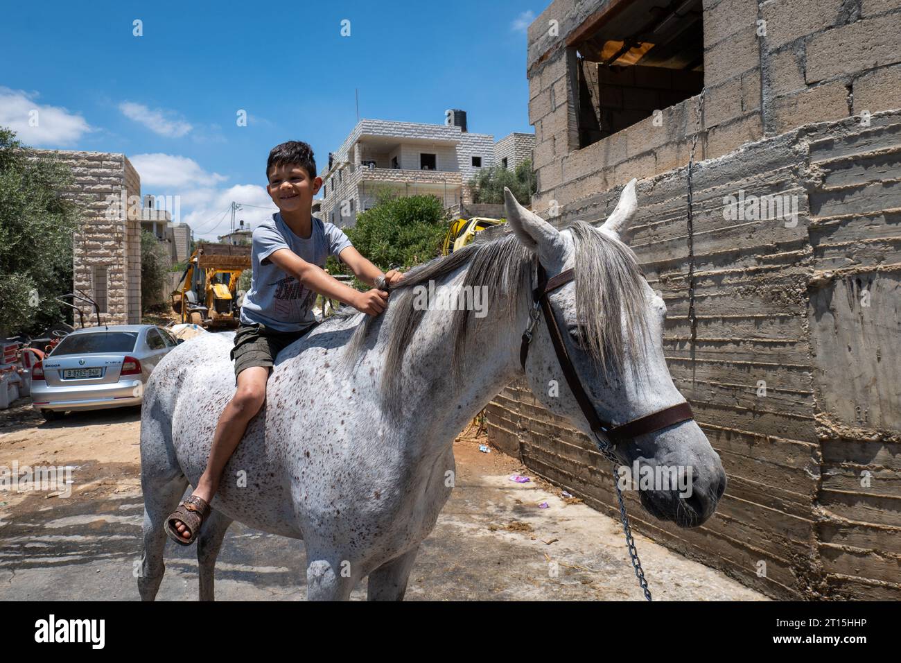 Bayt Surik, Palestina – 19 giugno 2023: Happy Young Arab Boy cavalca un bellissimo cavallo bianco e sorride in un edificio di cemento Foto Stock