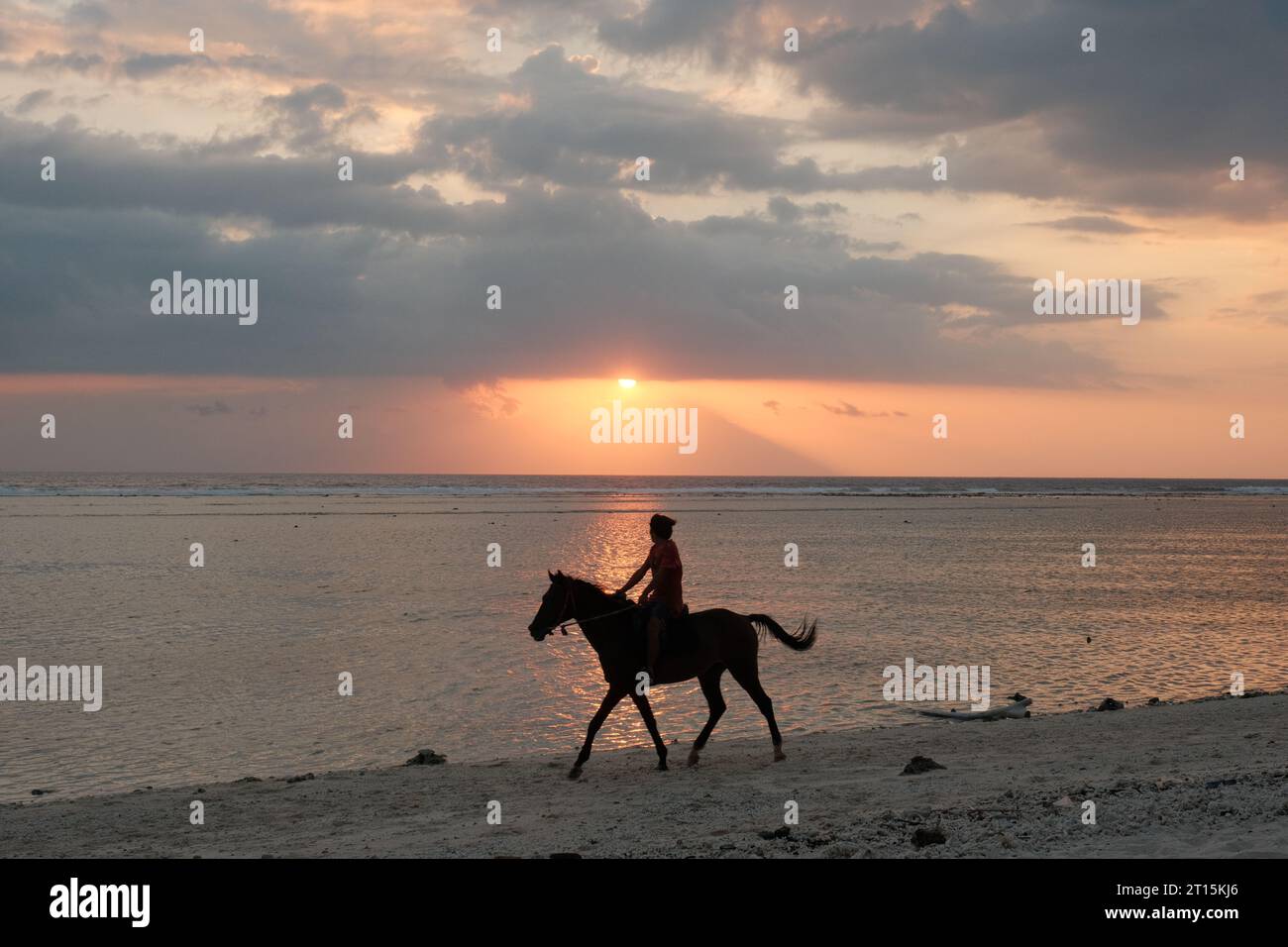 Un allineamento armonioso: Un uomo e il suo cavallo cavalcano lungo la linea verticale dove il tramonto bacia la spiaggia e il mare sul Gili Trawangan. Foto Stock