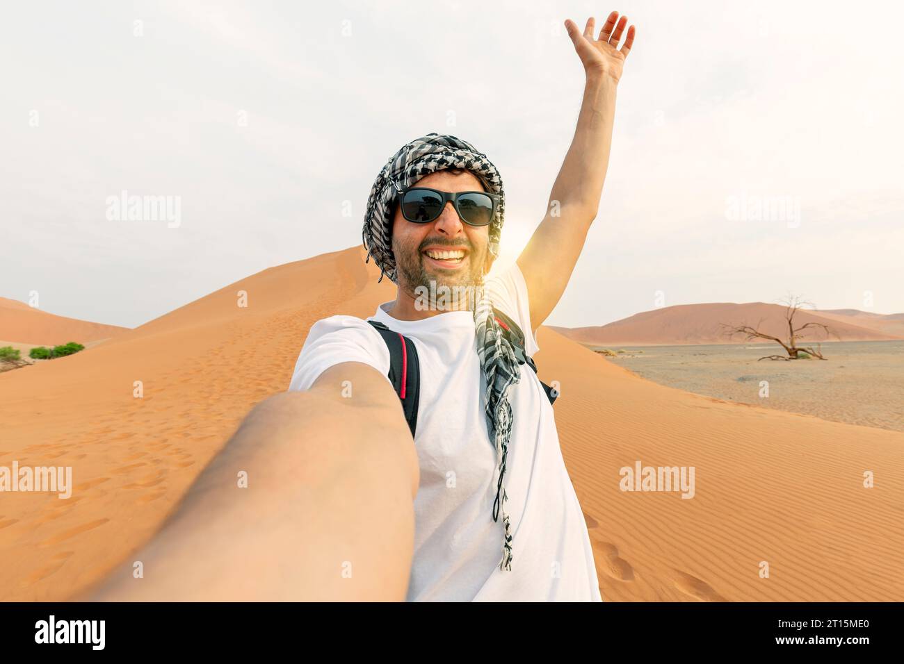 Giovane uomo con maglietta bianca nel deserto, che cammina negli Emirati Arabi Uniti durante il safari, indossa cappello e zaino, esplora la natura della bellezza sabbiosa. fare selfie dietro Foto Stock