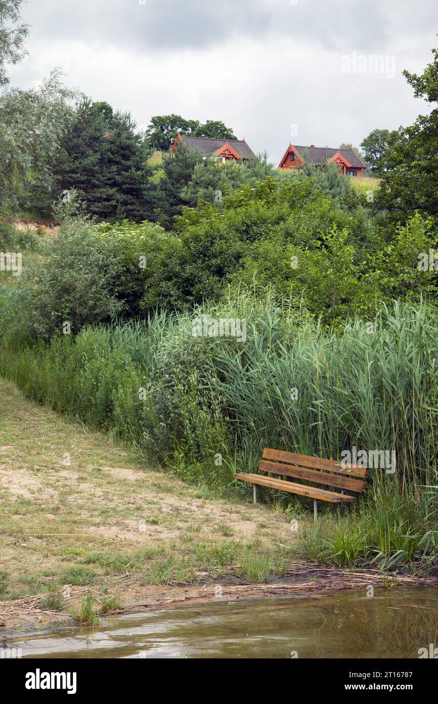 Una panchina di legno sulla riva di un lago con cottage di paglia su una collina Foto Stock