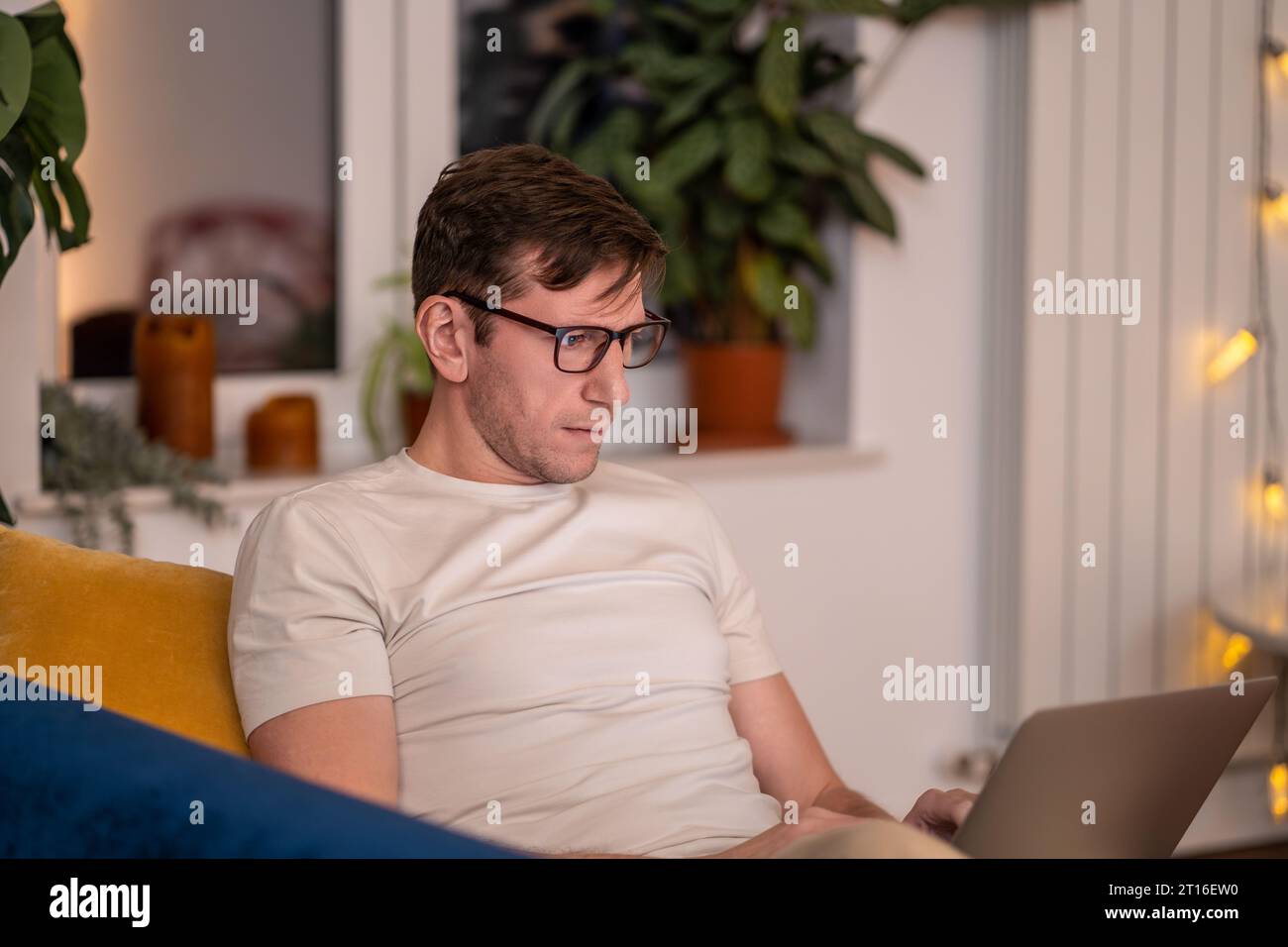 Un uomo seduto sul divano con un computer portatile sul grembo fa gli straordinari. Internet remoto online per studiare lavoro da casa Foto Stock