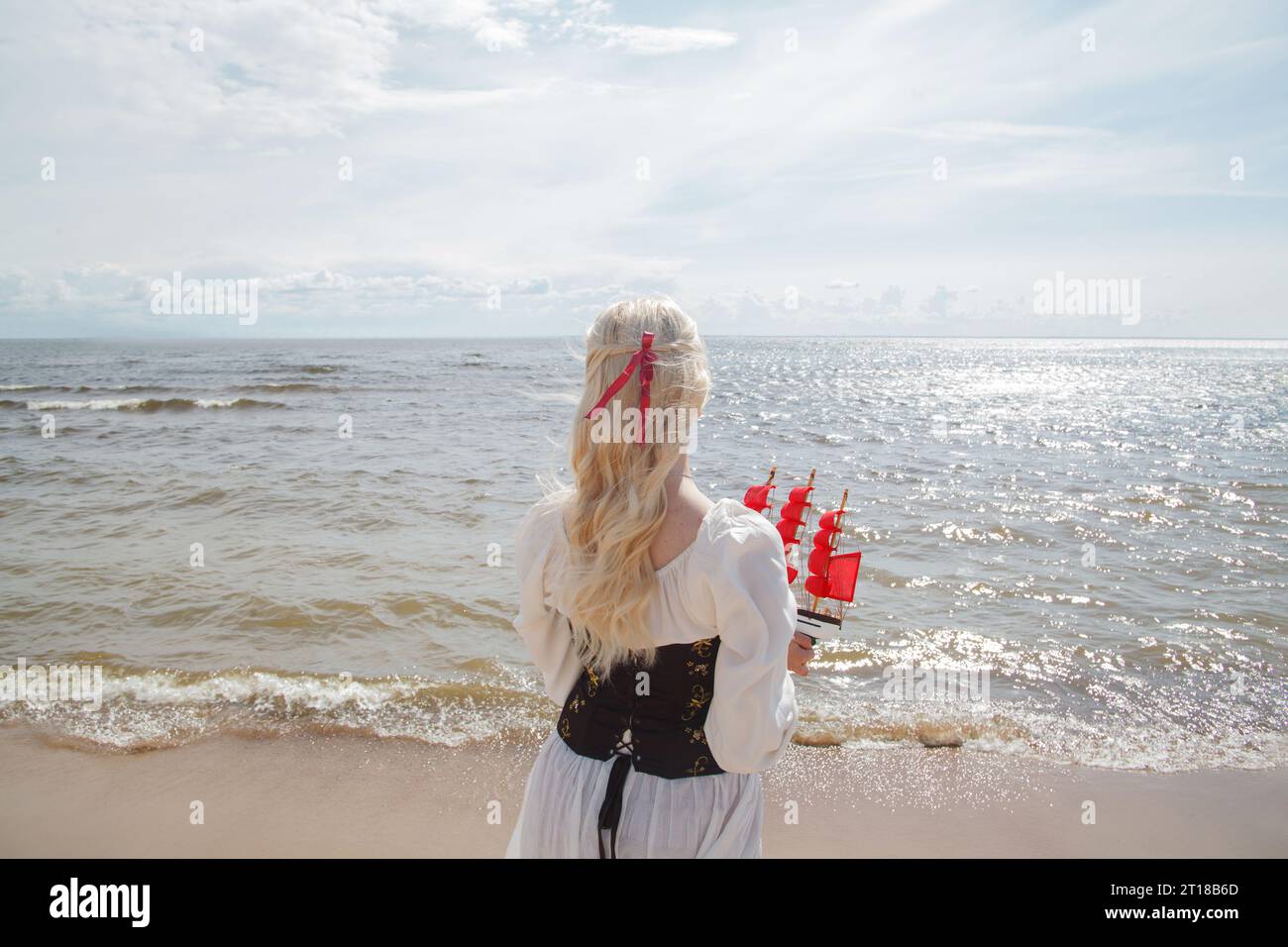 Giovane donna bionda romantica in piedi sulla spiaggia di mare con vele rosse piccola barca, ritratto all'aperto Foto Stock