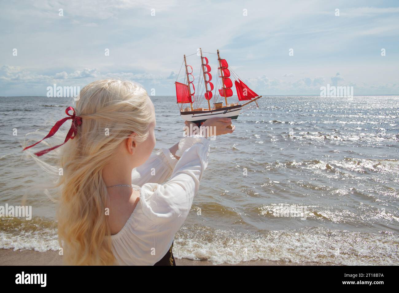 Giovane donna bionda che regge un piccolo yacht con vele rosse sulla spiaggia di mare Foto Stock