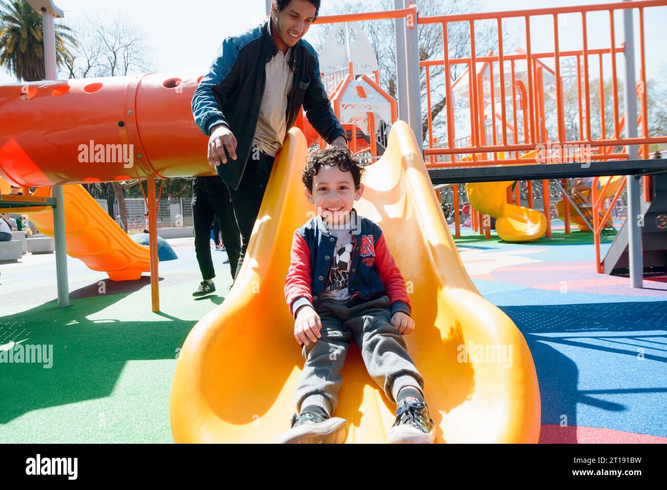 giovane latinoamericano felice padre single che gioca con suo figlio sullo scivolo nel parco pubblico a mezzogiorno, condividendo con la famiglia nel fine settimana Foto Stock