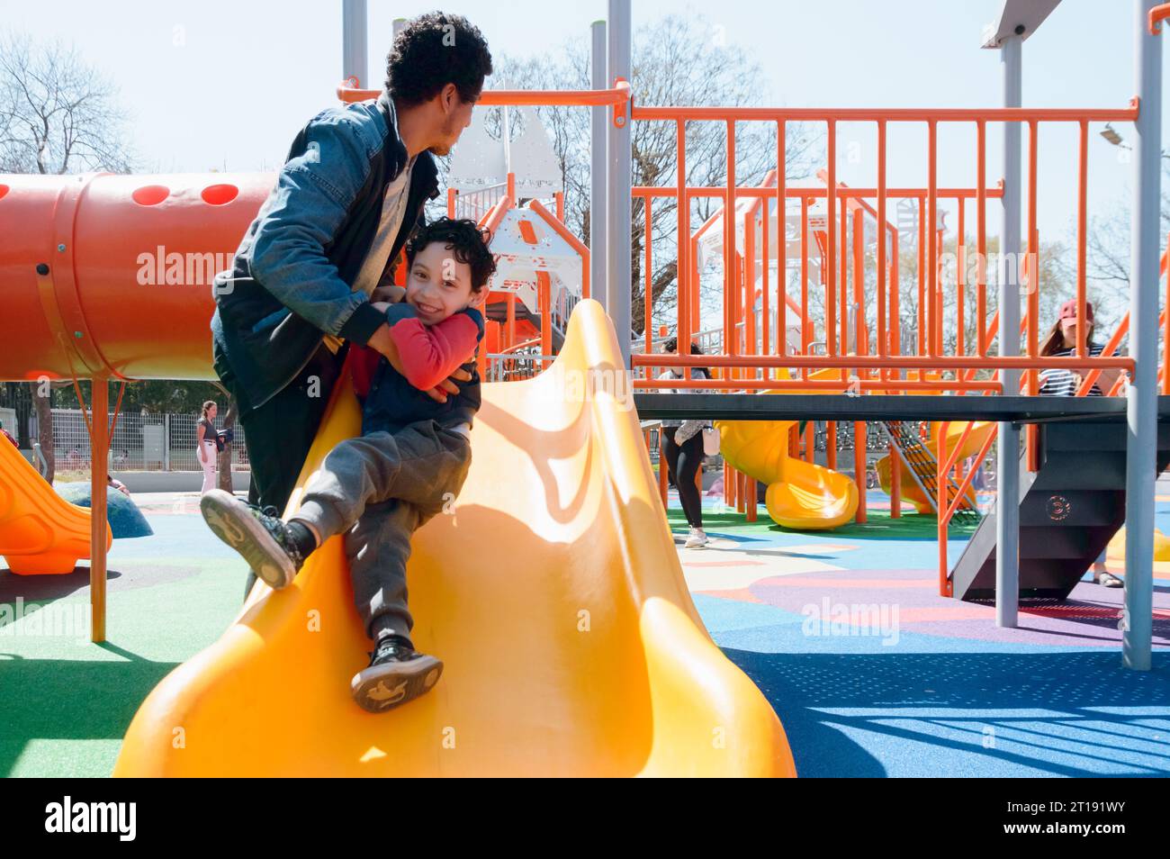 giovane latinoamericano felice padre single che gioca con suo figlio sullo scivolo nel parco pubblico a mezzogiorno, condividendo con la famiglia nel fine settimana Foto Stock