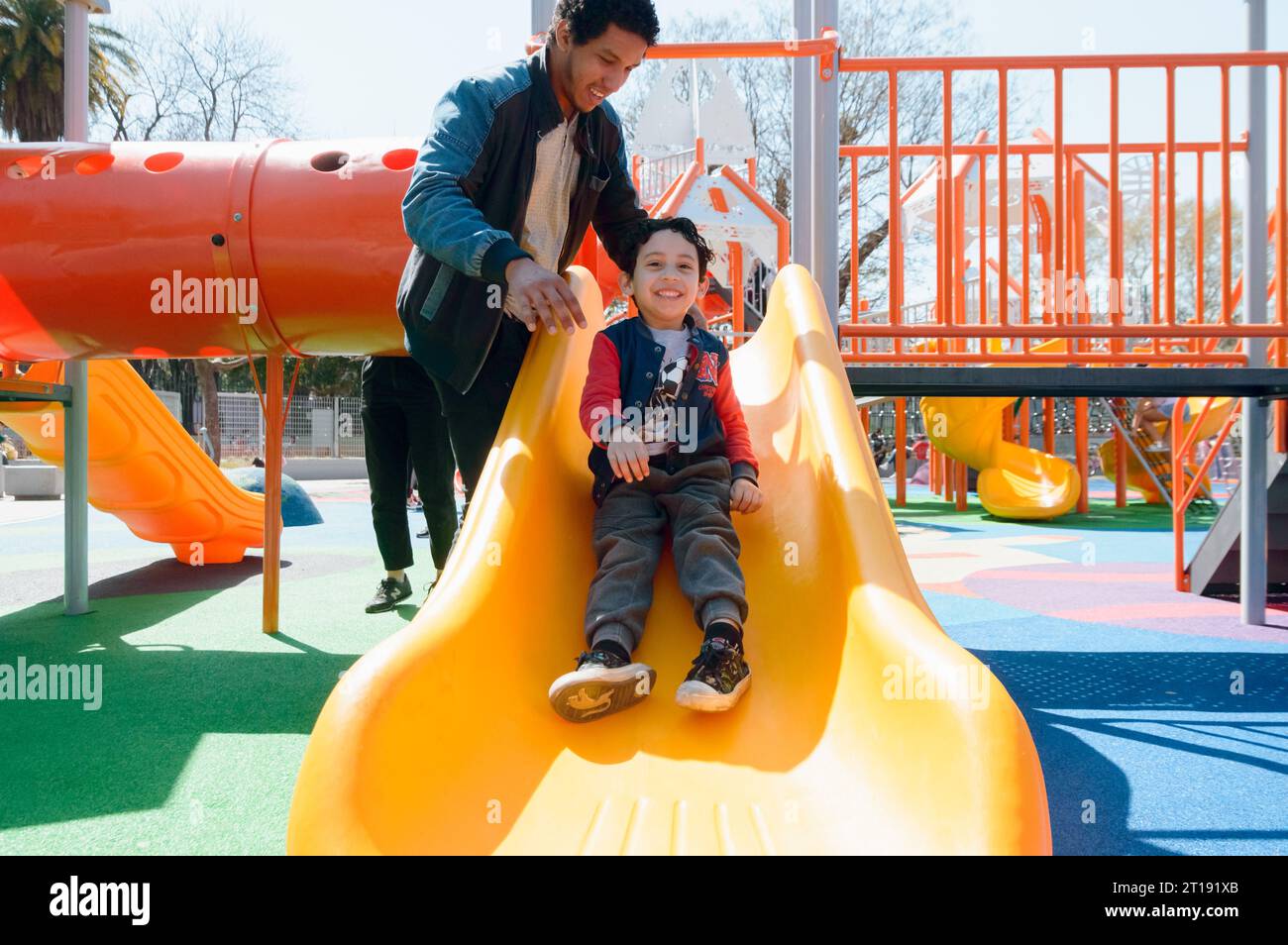 giovane latinoamericano felice padre single che gioca con suo figlio sullo scivolo nel parco pubblico a mezzogiorno, condividendo con la famiglia nel fine settimana Foto Stock