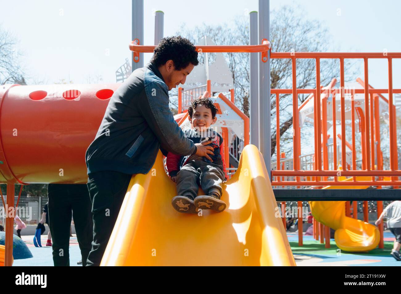 giovane latinoamericano felice padre single che gioca con suo figlio sullo scivolo nel parco pubblico a mezzogiorno, condividendo con la famiglia nel fine settimana Foto Stock