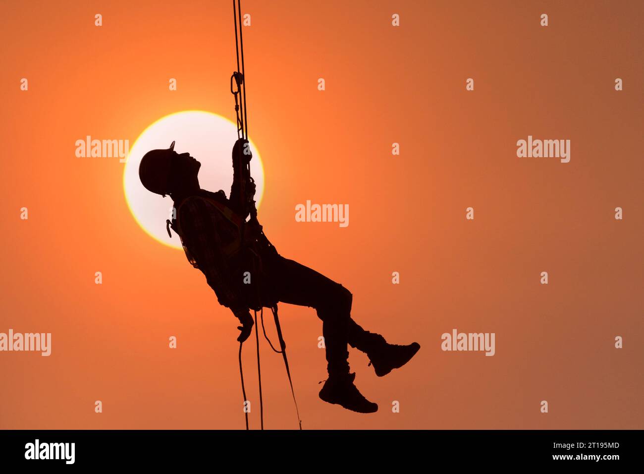 Silhouette di un operaio edile appeso a un'imbracatura di sicurezza contro un cielo arancione al tramonto, Thailandia Foto Stock