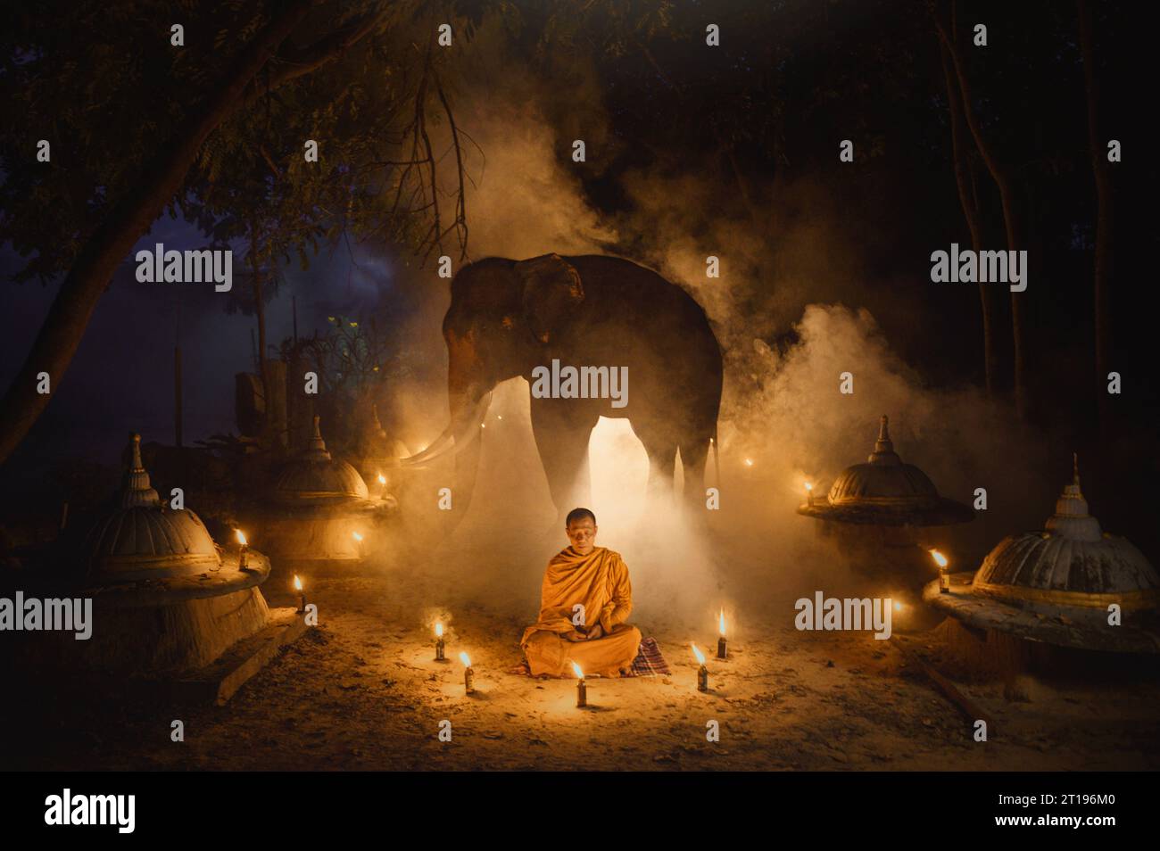 Monaco in un tradizionale abito arancio (kasay) circondato da candele seduto di fronte ad un elefante, Surin, Thailandia Foto Stock