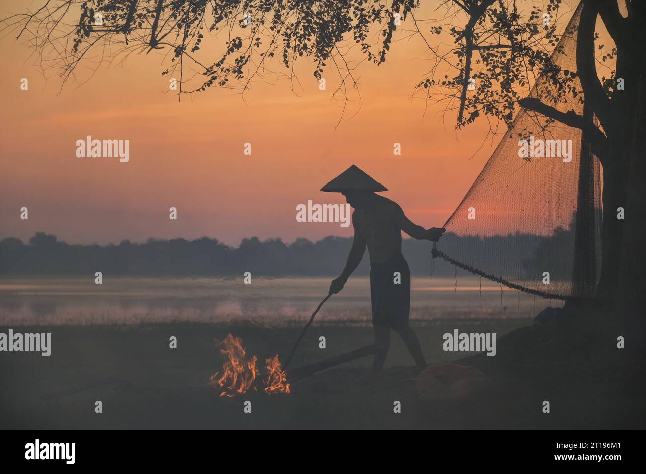 Sagoma di un pescatore che tiene una rete da pesca e fa un incendio sulla riva del fiume al tramonto, Thailandia Foto Stock