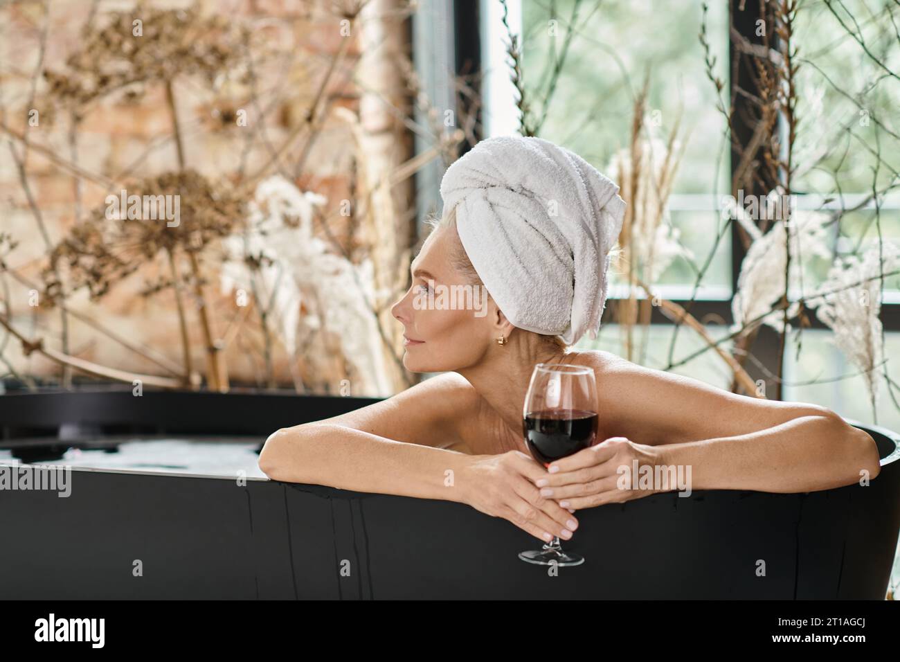 donna rilassata e sognante con un asciugamano bianco sulla testa che regge un bicchiere di vino rosso e fa il bagno Foto Stock