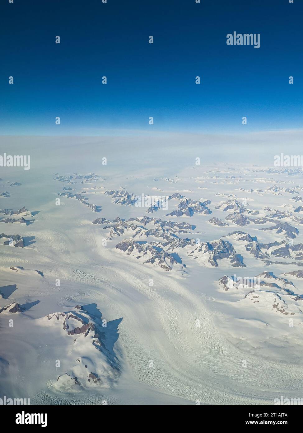 Vista dall'alto da un piano di estesi campi di neve e cime montuose della Groenlandia in una giornata di sole con cielo blu all'orizzonte Foto Stock