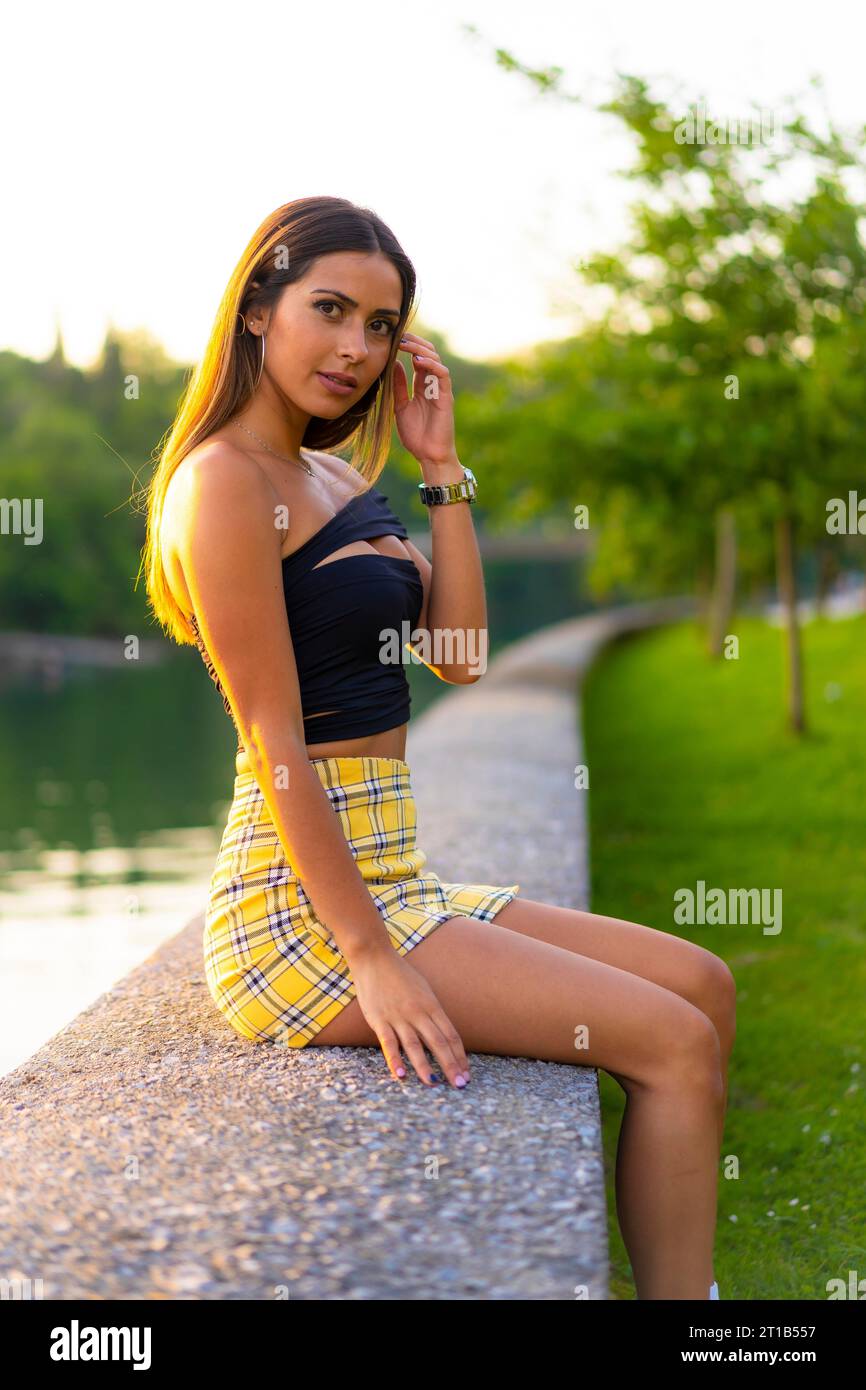 Lifestyle, una graziosa giovane donna bruna caucasica con il top nero stretto e la gonna gialla, seduta in un verde parco cittadino al tramonto Foto Stock