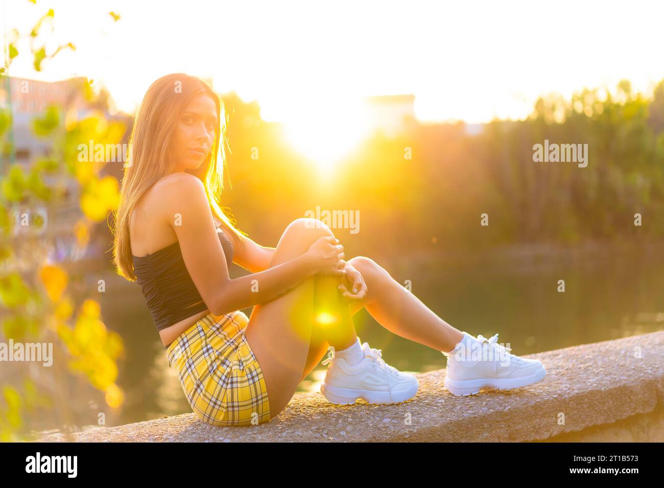 Lifestyle, una giovane donna seduta al tramonto in un parco, donna caucasica dai capelli scuri con top nero stretto e gonna gialla Foto Stock