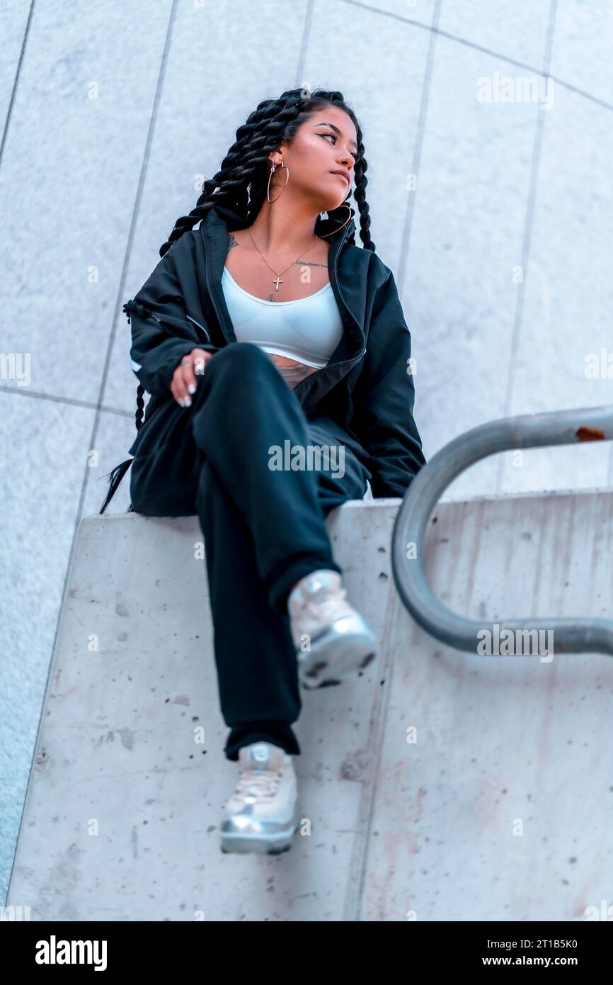 Sessione urbana. Giovane donna di etnia nera con lunghe trecce e tatuaggi, seduta in città Foto Stock
