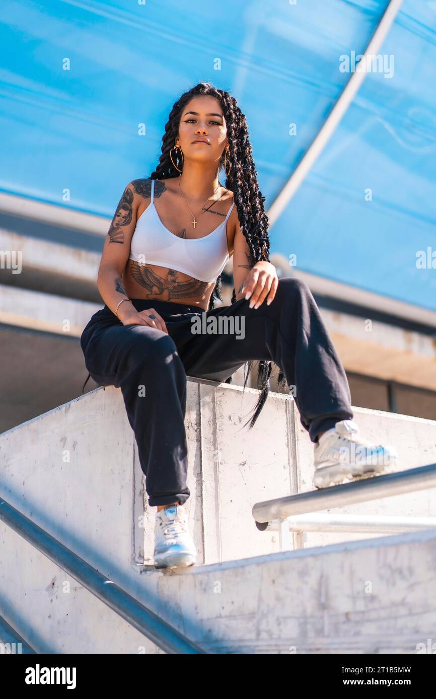 Giovane donna di etnia nera con lunghe trecce e tatuaggi, seduta sul cemento, Urban Shoot Foto Stock