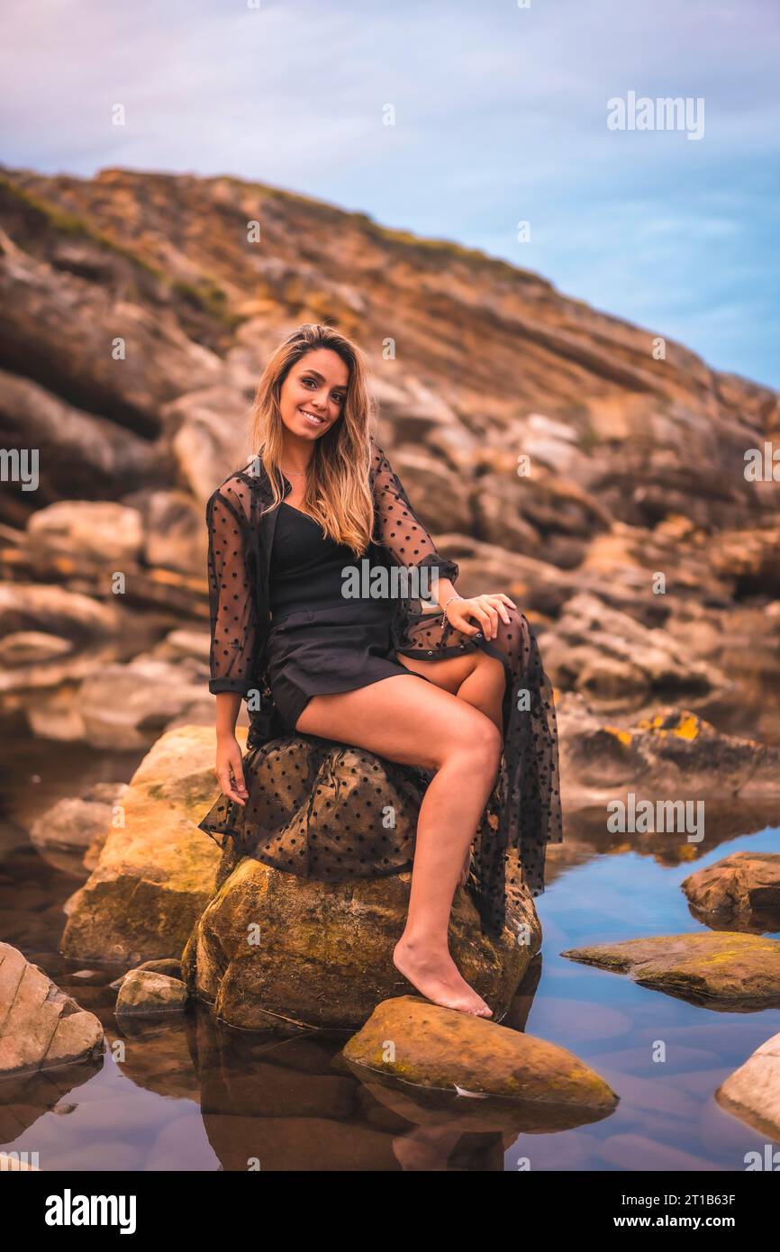 Stile di vita estivo con una giovane bruna caucasica in un lungo abito nero trasparente su alcune rocce vicino al mare in un pomeriggio d'estate. Seduto Foto Stock