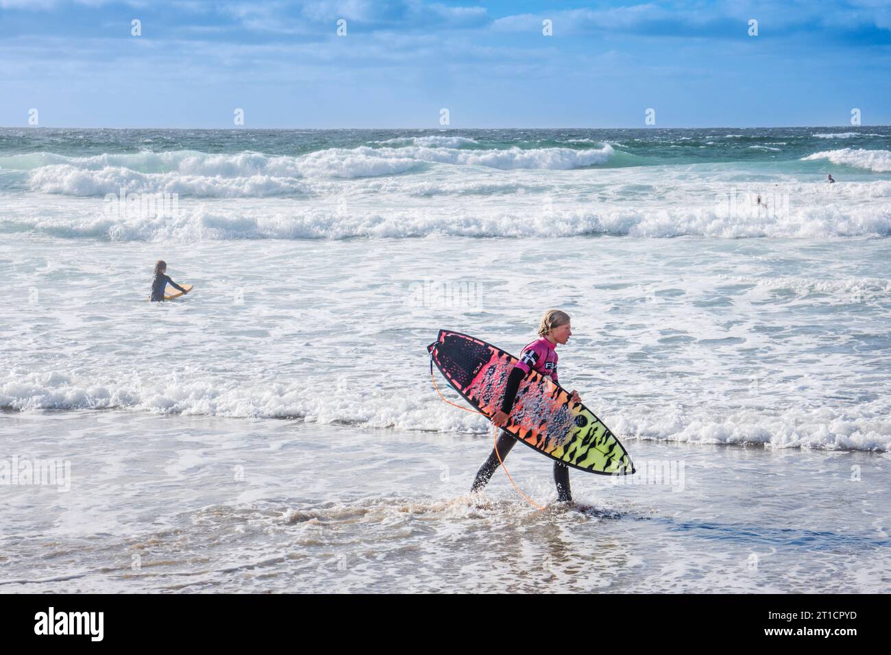 Un giovane surfista Grom che porta la sua tavola da surf a piedi lungo il litorale sull'iconica Fistral Beach a Newquay, in Cornovaglia, nel Regno Unito. Foto Stock