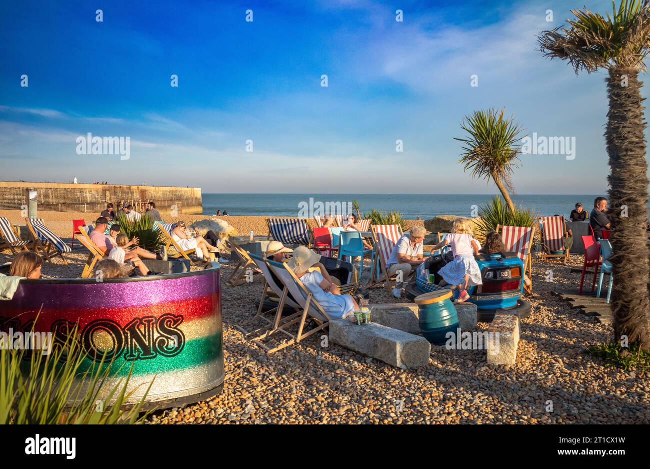 Le persone si siedono su sdraio in una giornata di sole sulla spiaggia al Pilot Beach Bar di Folkestone, Kent, Regno Unito. Foto Stock