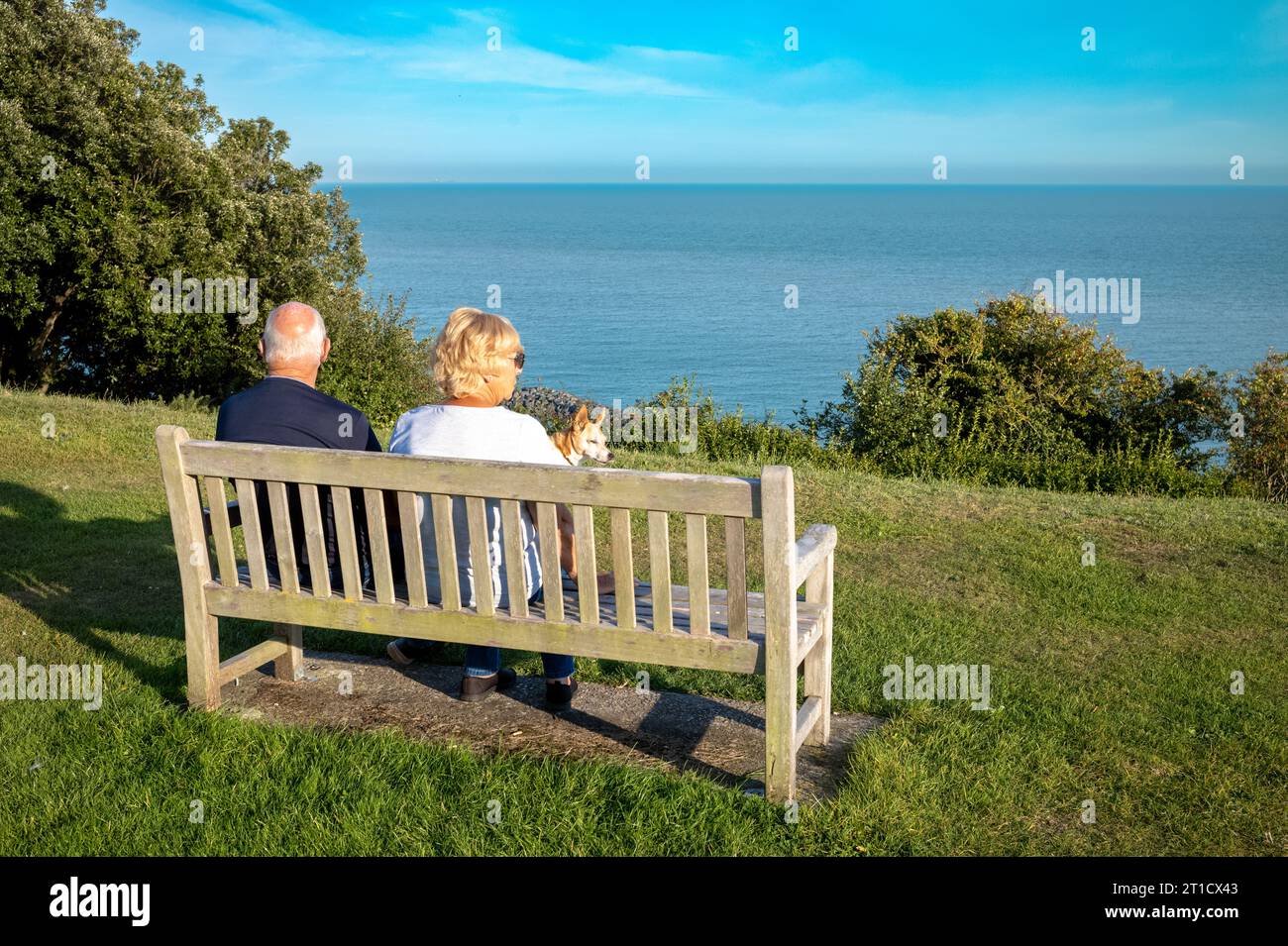 Una coppia di anziani in pensione e il loro cane si siedono su una panchina pubblica e guardano il Canale della Manica a Folkestone, Kent, Regno Unito. Foto Stock